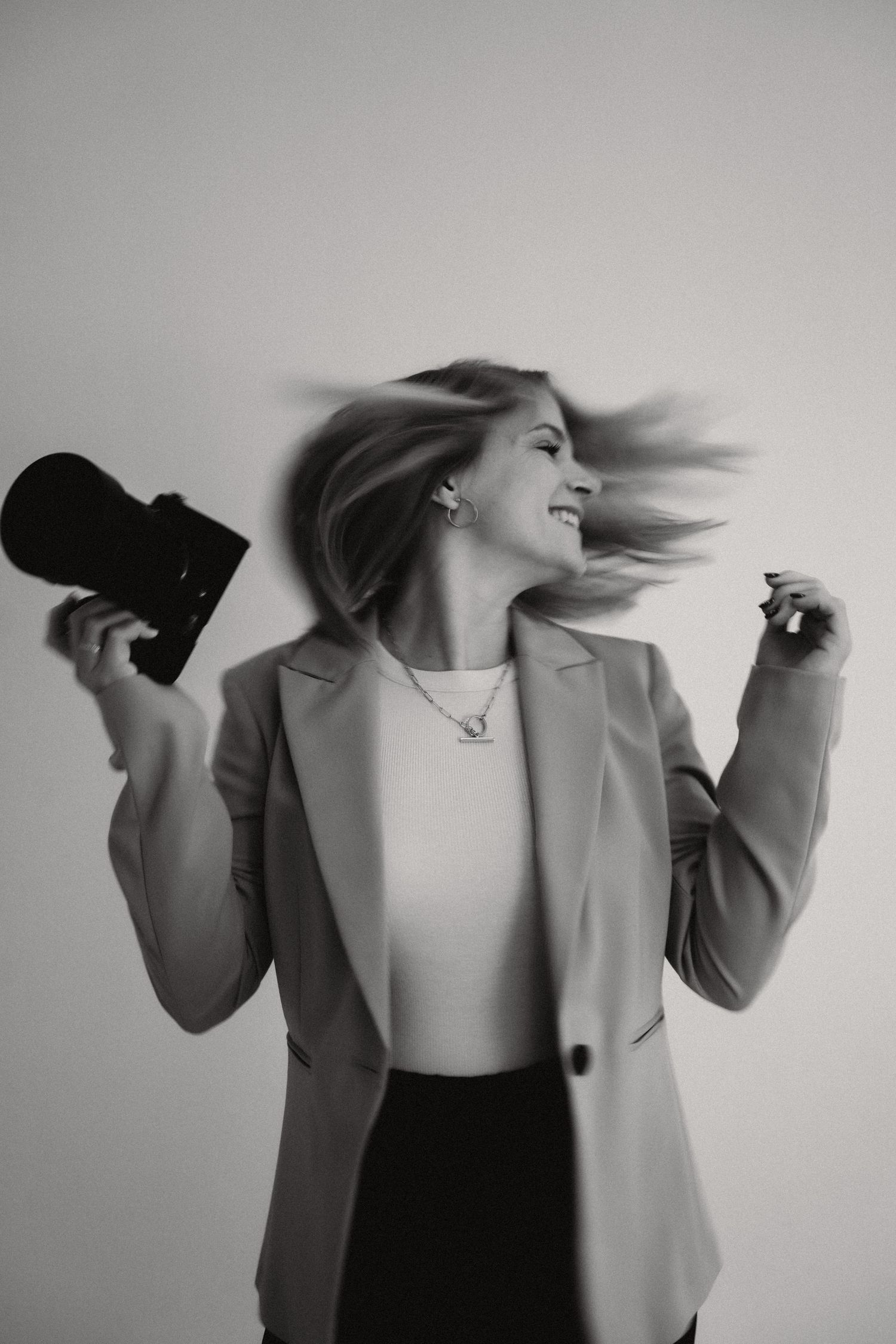 Black and white portrait of someone in a blazer holding a camera, with hair flowing in motion against light background.