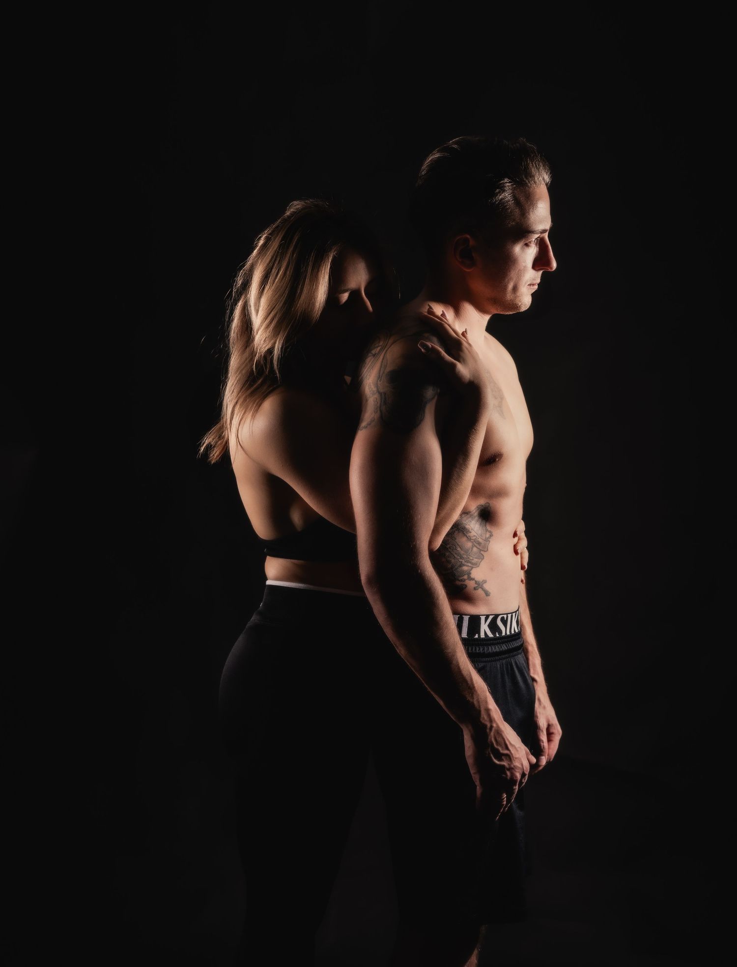 Fit couple posing back-to-back in dark studio, shirtless man with tattoos and woman in sports crop top.