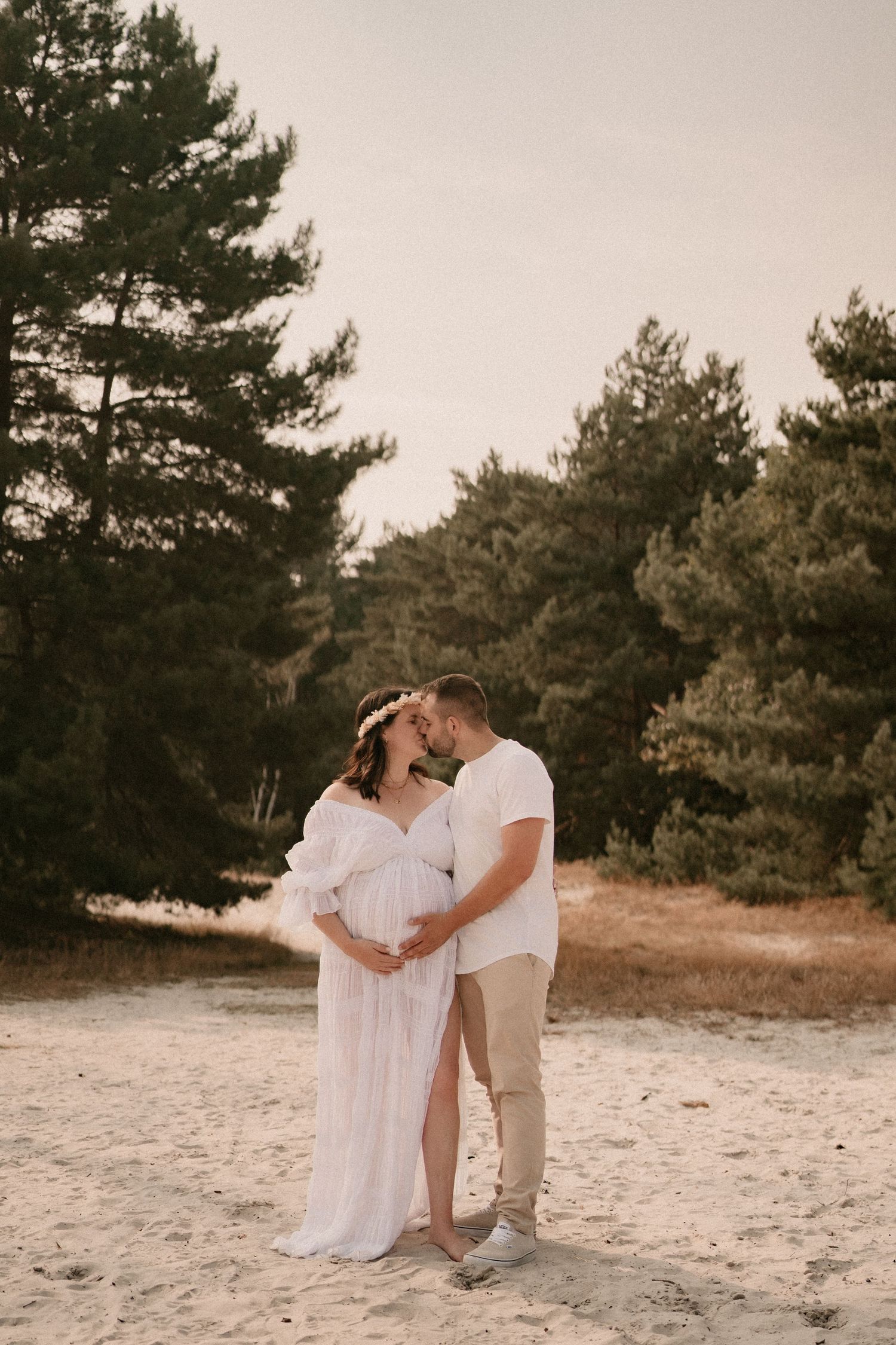 A romantic couple in white and beige outfits share tender moments on a sandy beach near pine trees.
