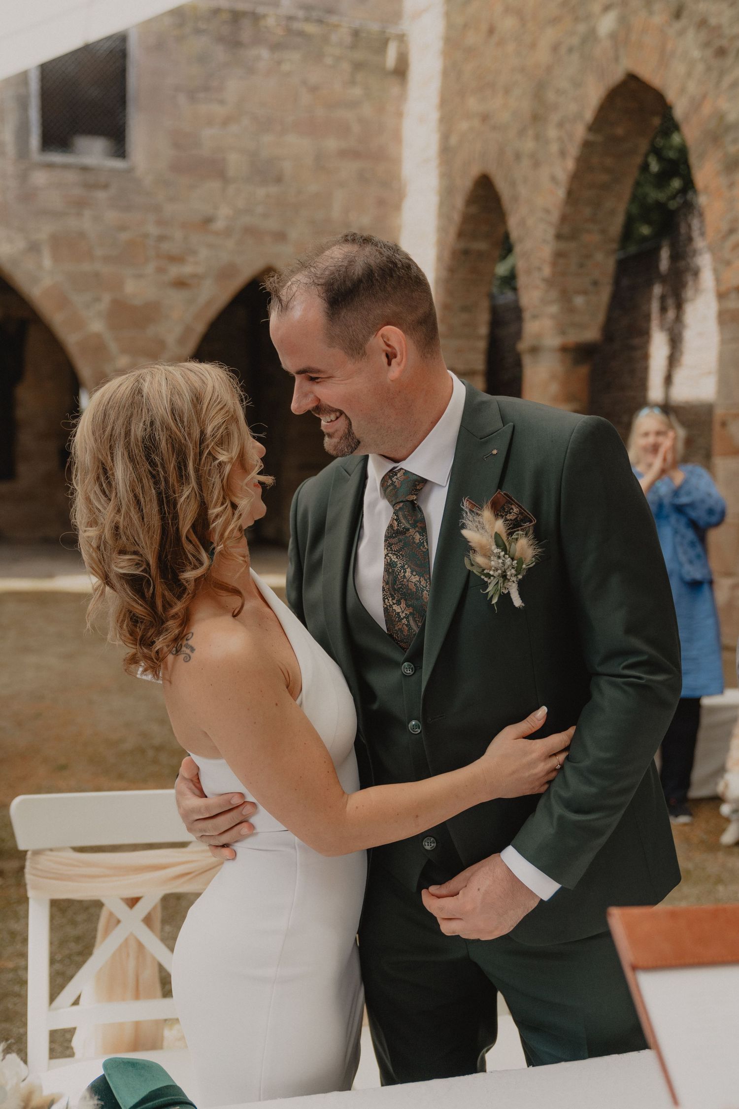 A romantic moment between newlyweds sharing a laugh at their wedding ceremony against a stone archway backdrop.