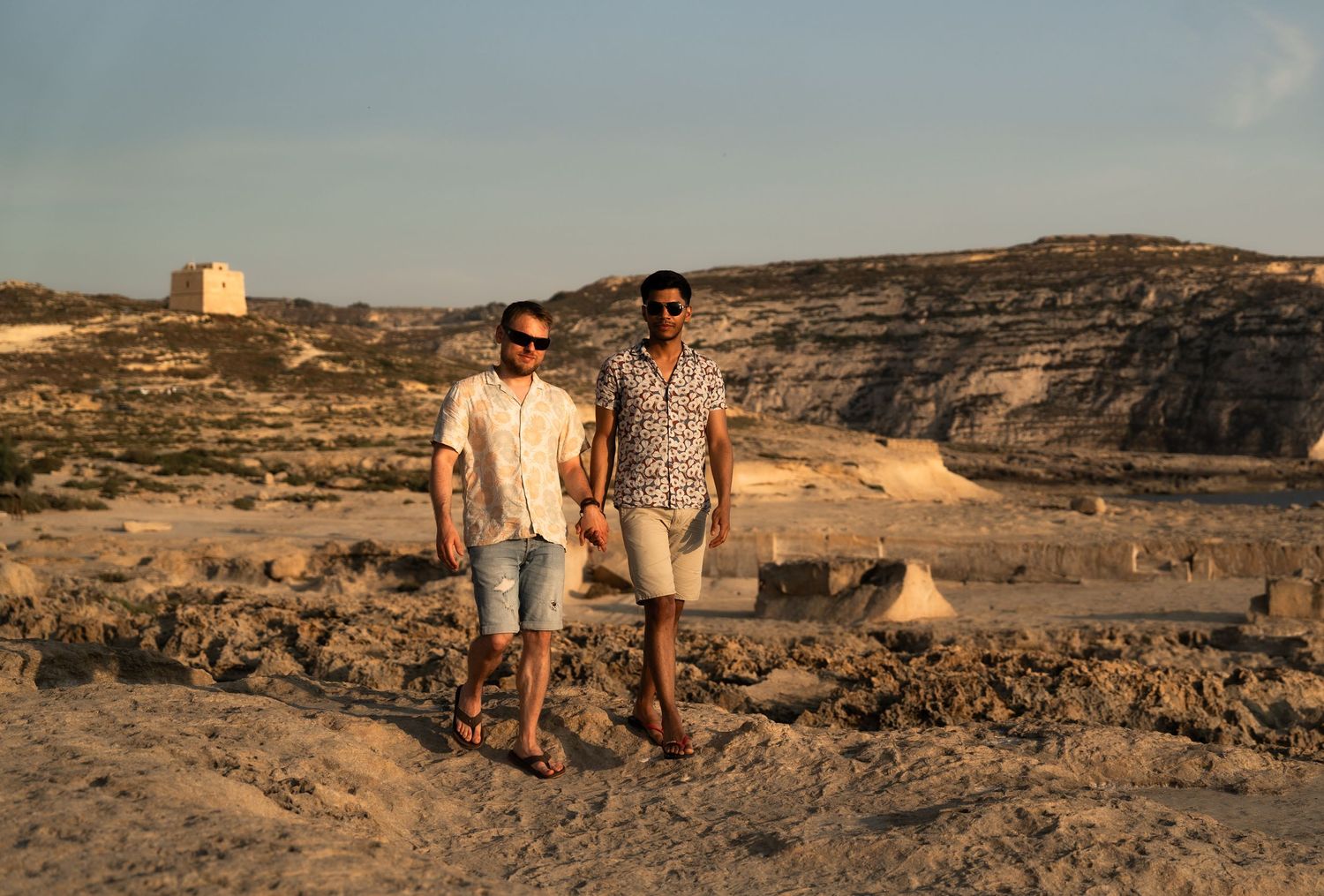 Two men walking hand in hand through a rocky desert landscape at golden hour, wearing casual summer outfits and sunglasses.