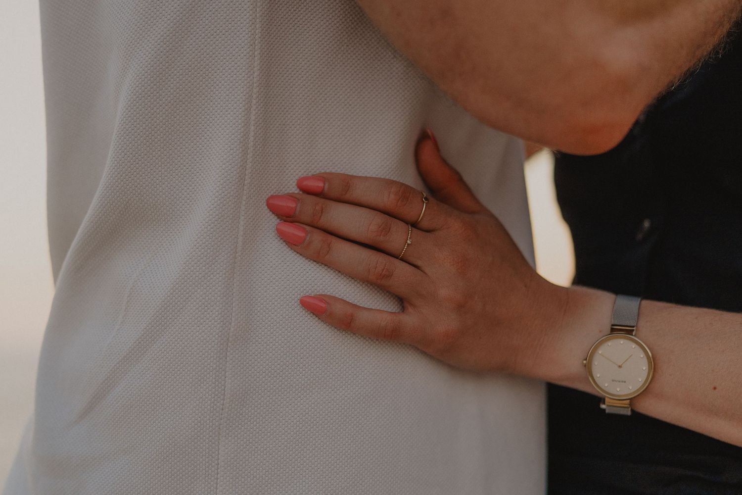 Close-up of a woman's hand with pink nails and gold watch resting on a man's chest in an embrace.