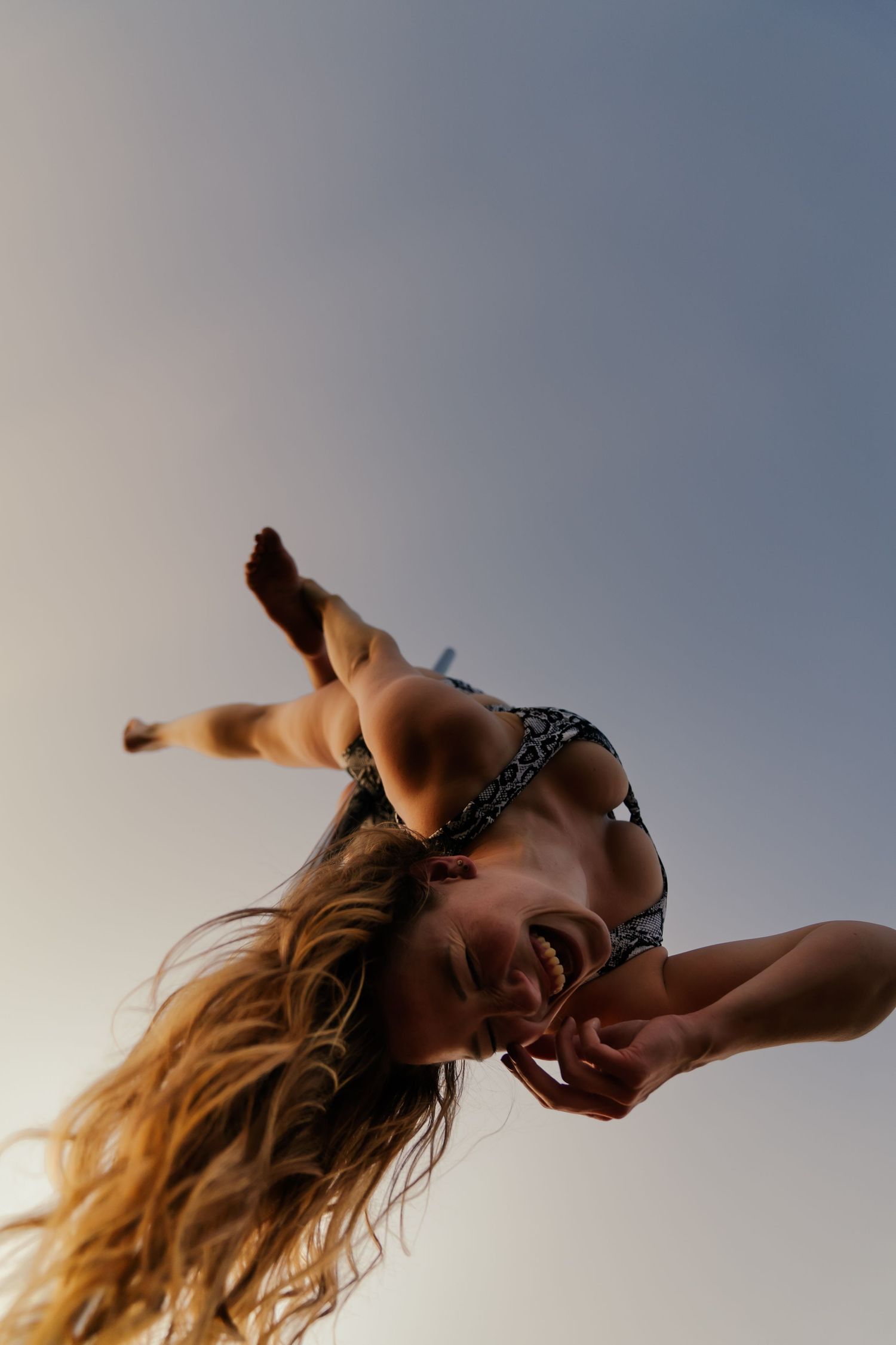 A young woman in a bikini laughs joyfully while jumping upside down against a clear sky backdrop.