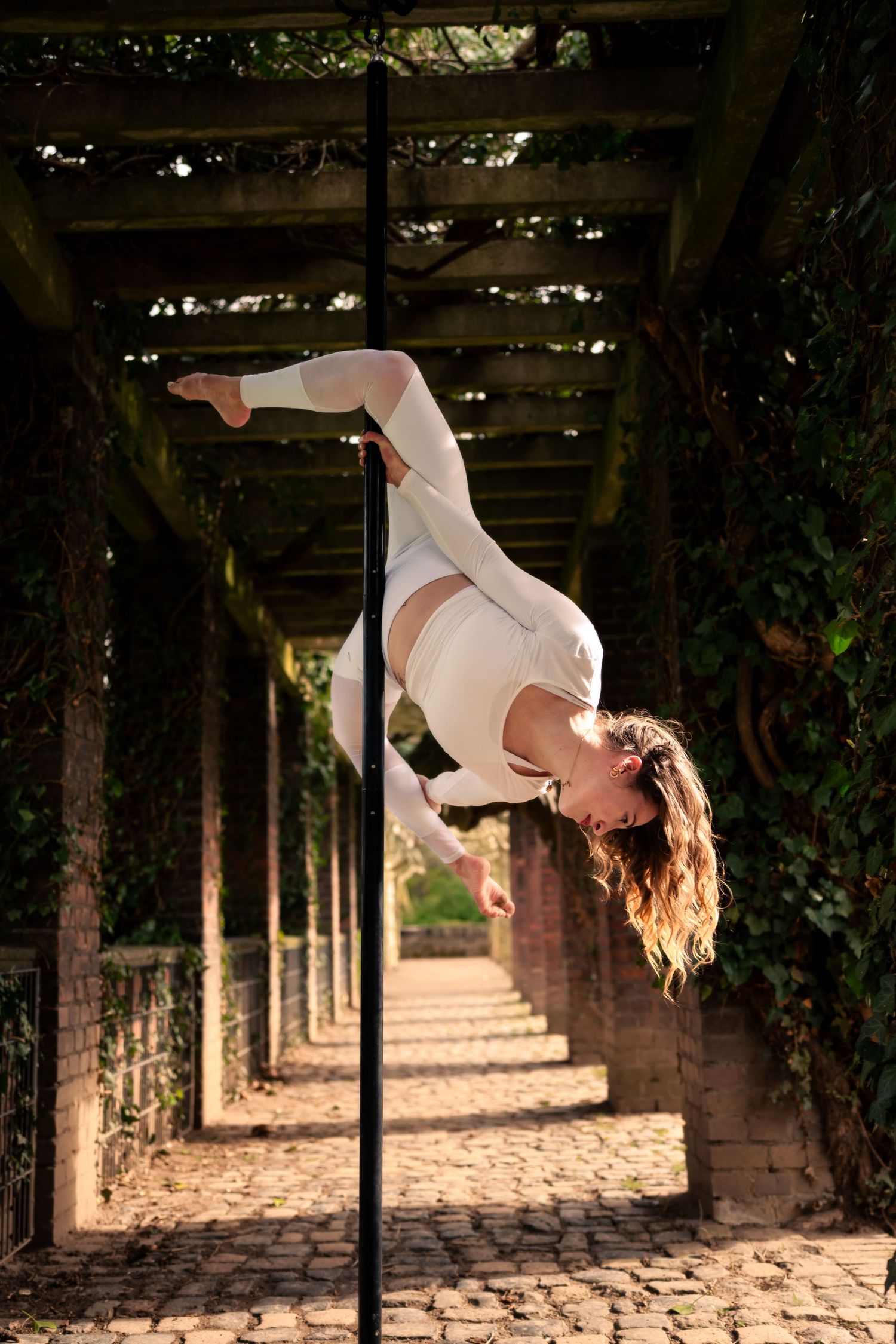 Woman in white outfit performing an inverted pole dance move outdoors in a vine-covered pergola walkway.