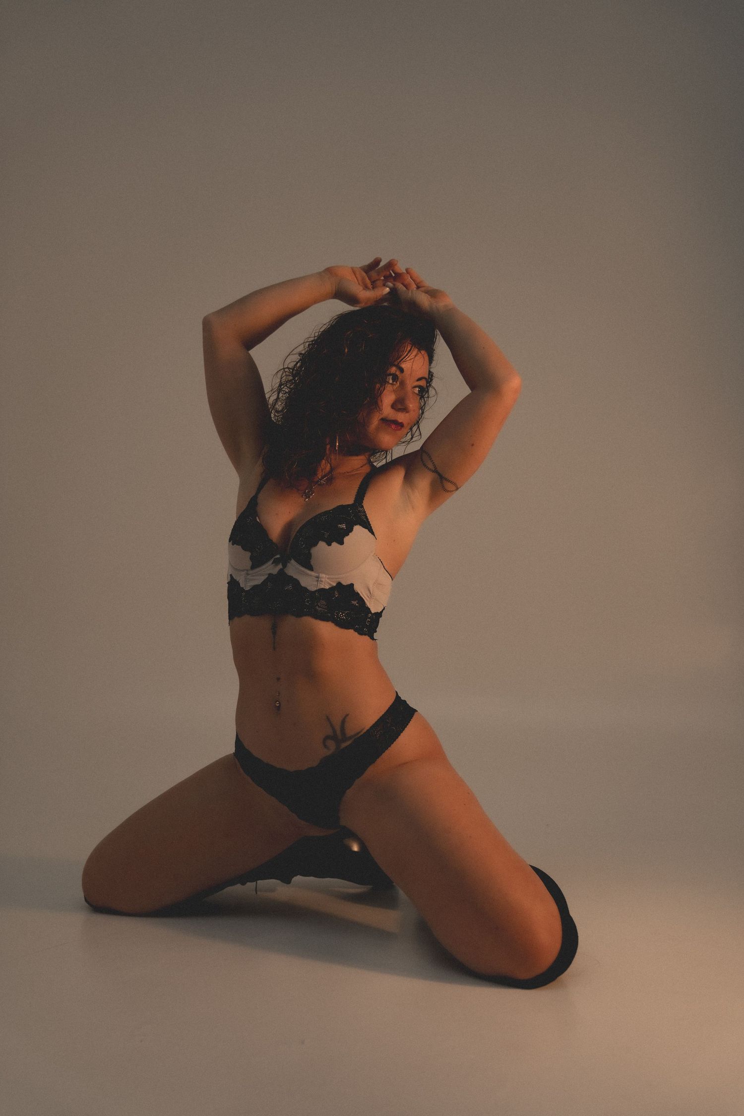 Woman in black lace lingerie posing on knees with arms raised, studio photography on gray background.