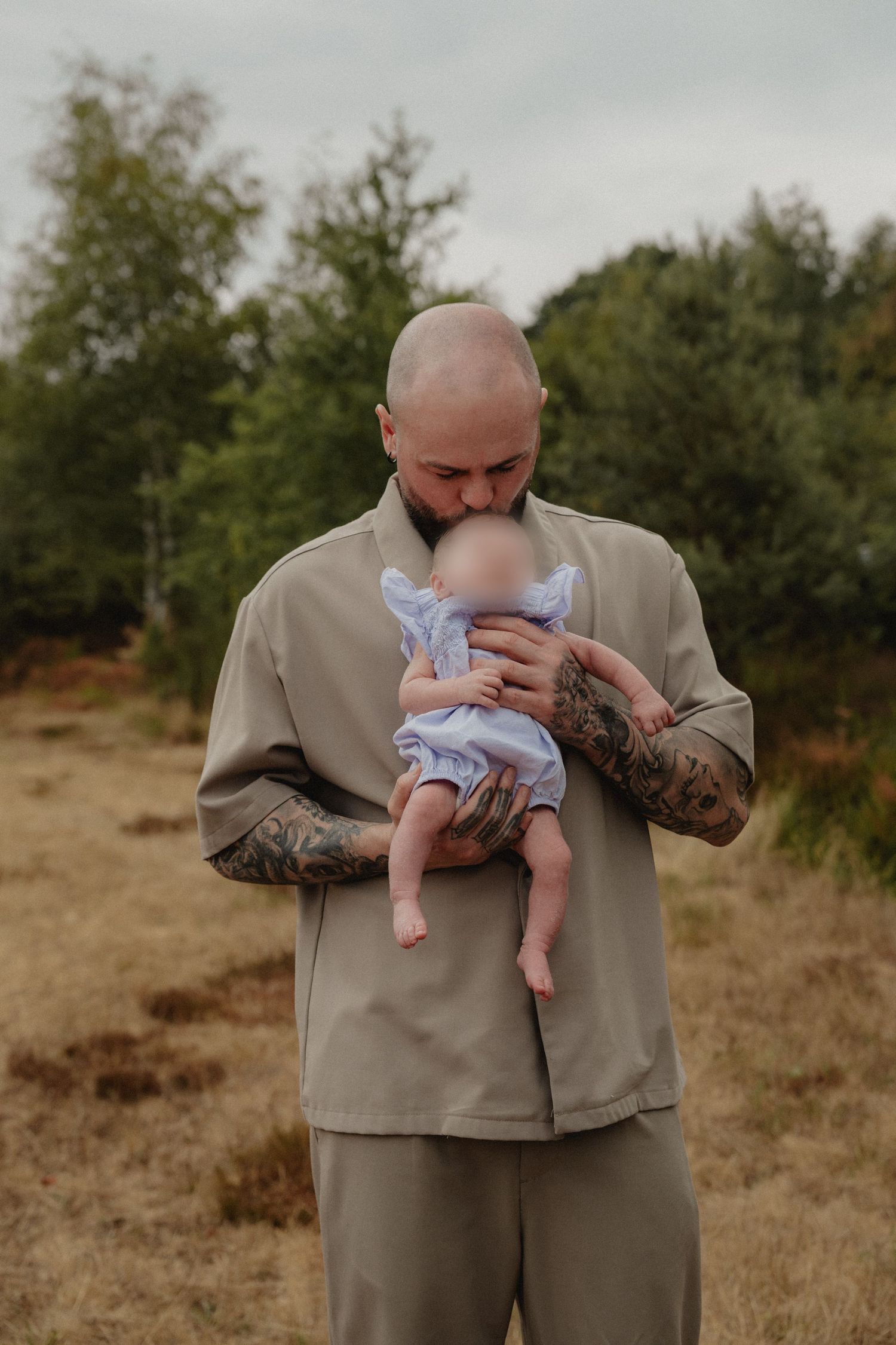 Tattooed bald man tenderly kissing baby in blue outfit outdoors in a field.