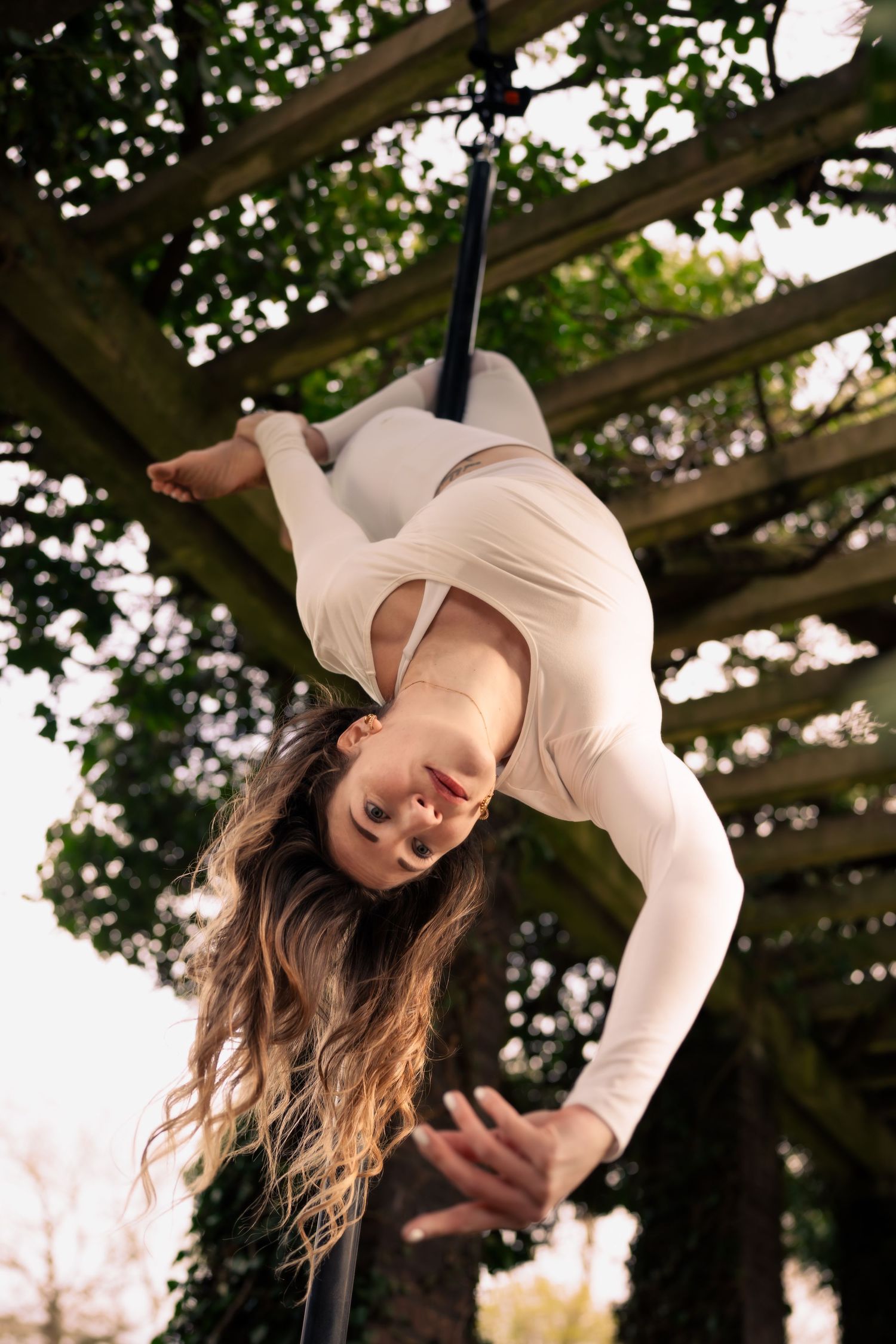 Woman in white outfit performing aerial acrobatics upside down on silks outdoors under wooden pergola.