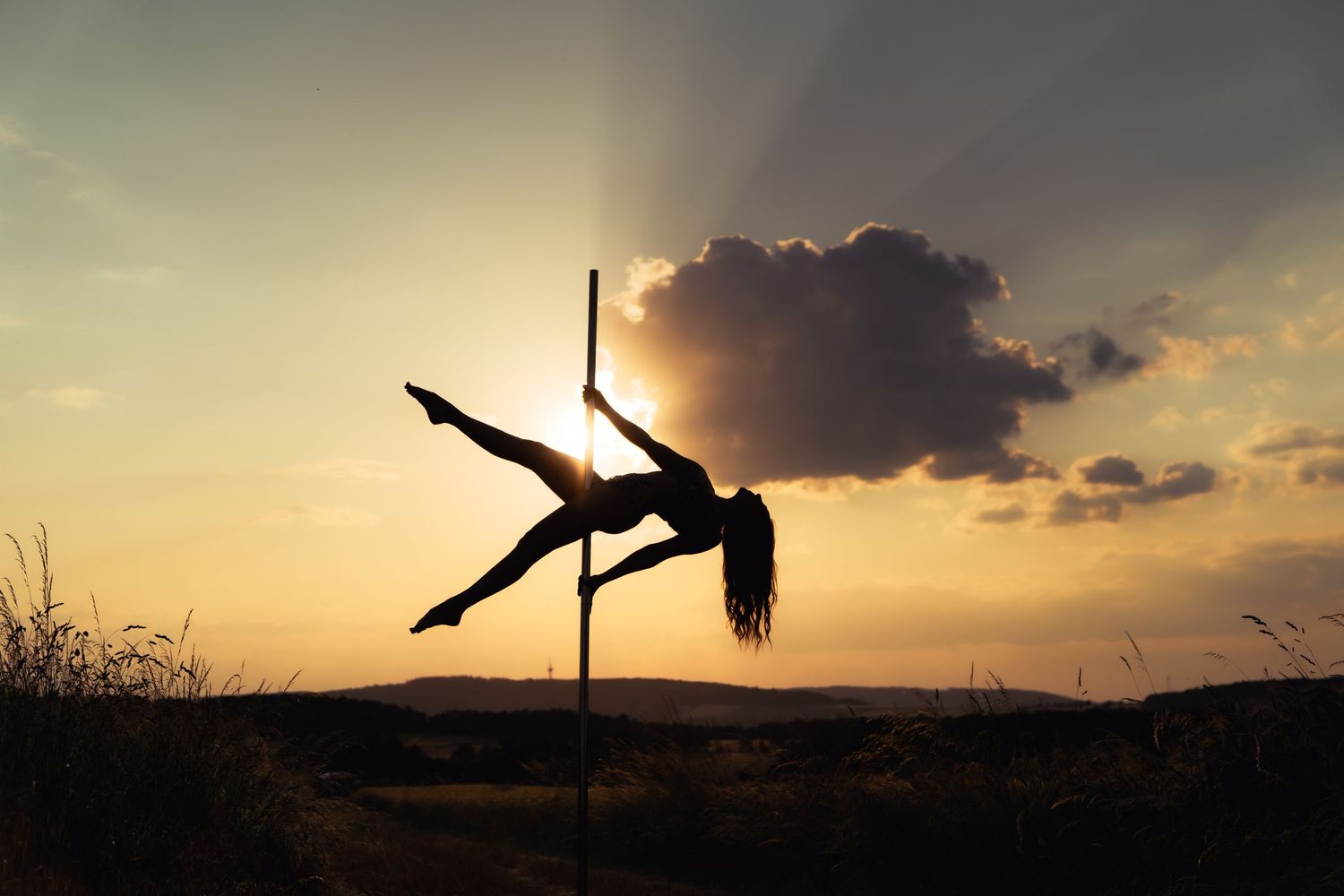 Silhouette of a woman pole dancing outdoors against a dramatic sunset sky with glowing clouds.