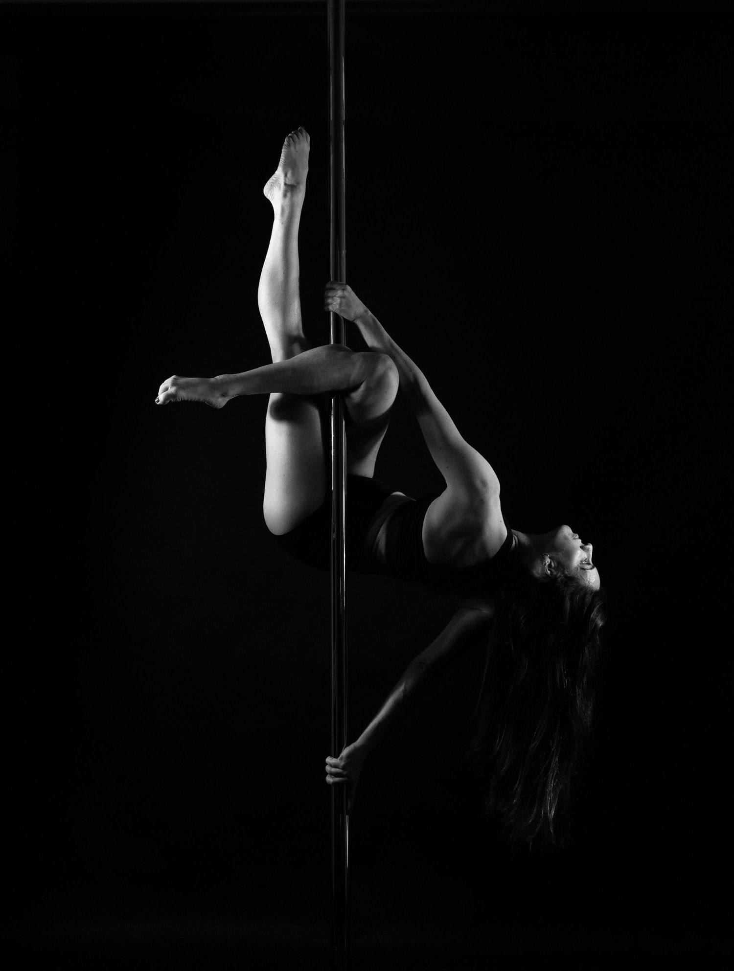 Black and white photo of a woman performing an impressive pole dance pose against a dark background.