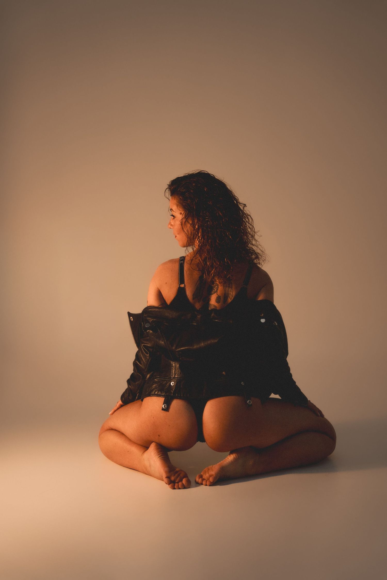 Woman with curly hair sitting cross-legged on floor in black outfit, bathed in warm golden studio light.