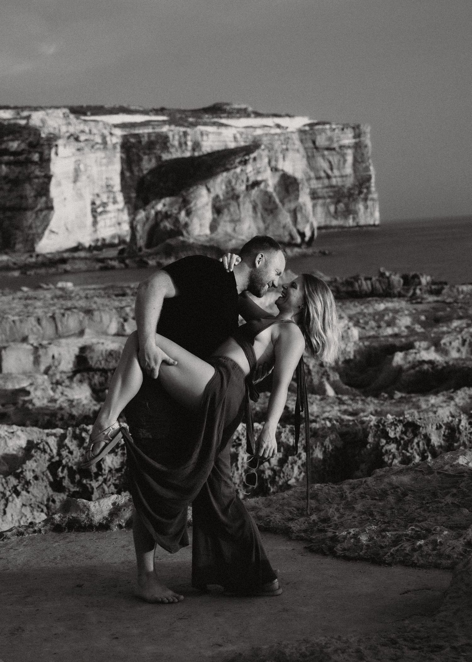 Black and white photo of a couple romantically embracing near coastal cliffs, man lifting woman in elegant dress.