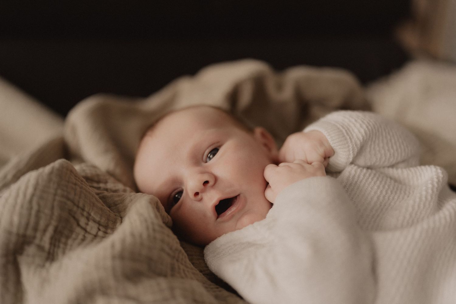 Newborn baby in white outfit resting on soft neutral bedding.