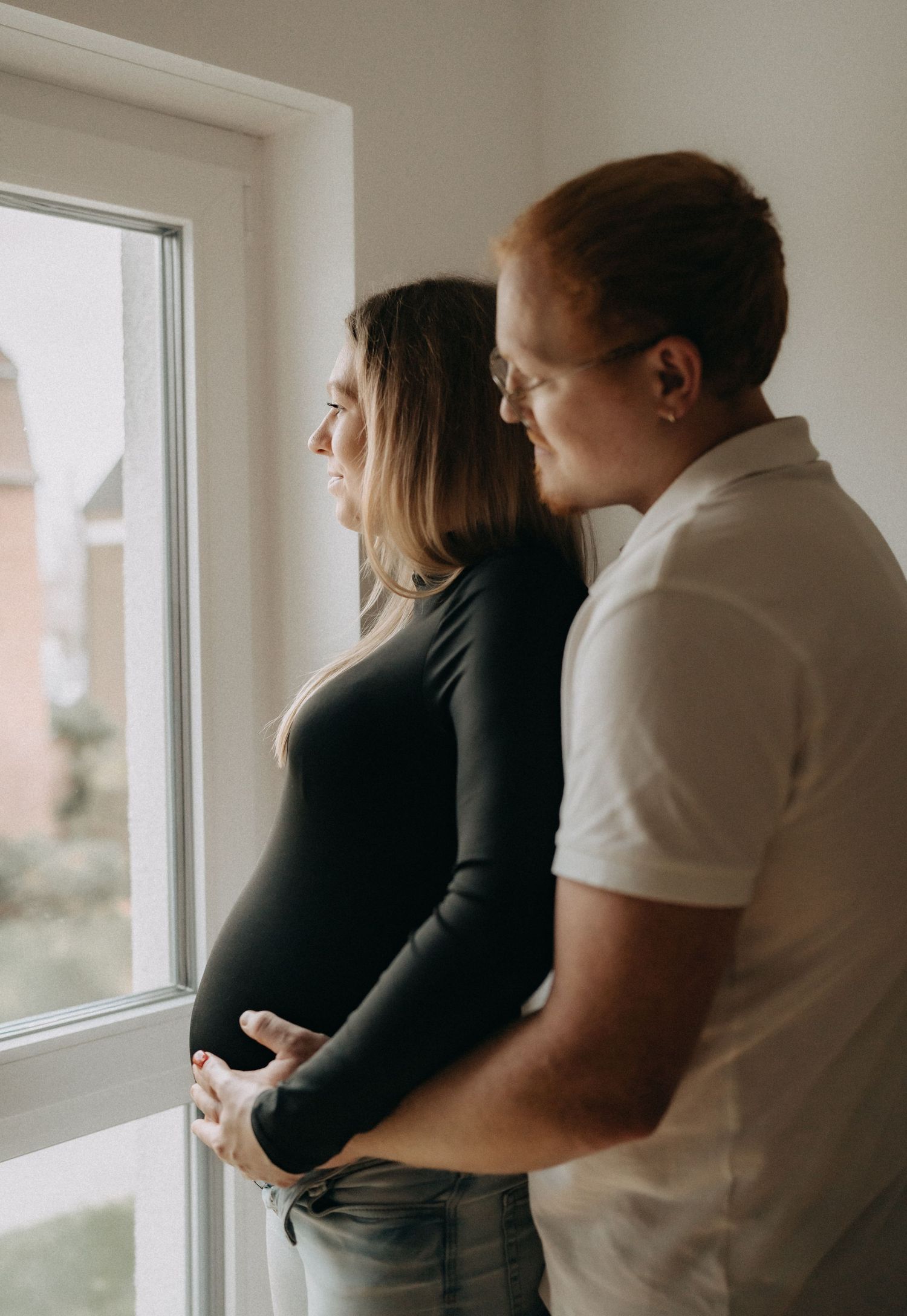A couple stands by a window sharing an intimate pregnancy moment.