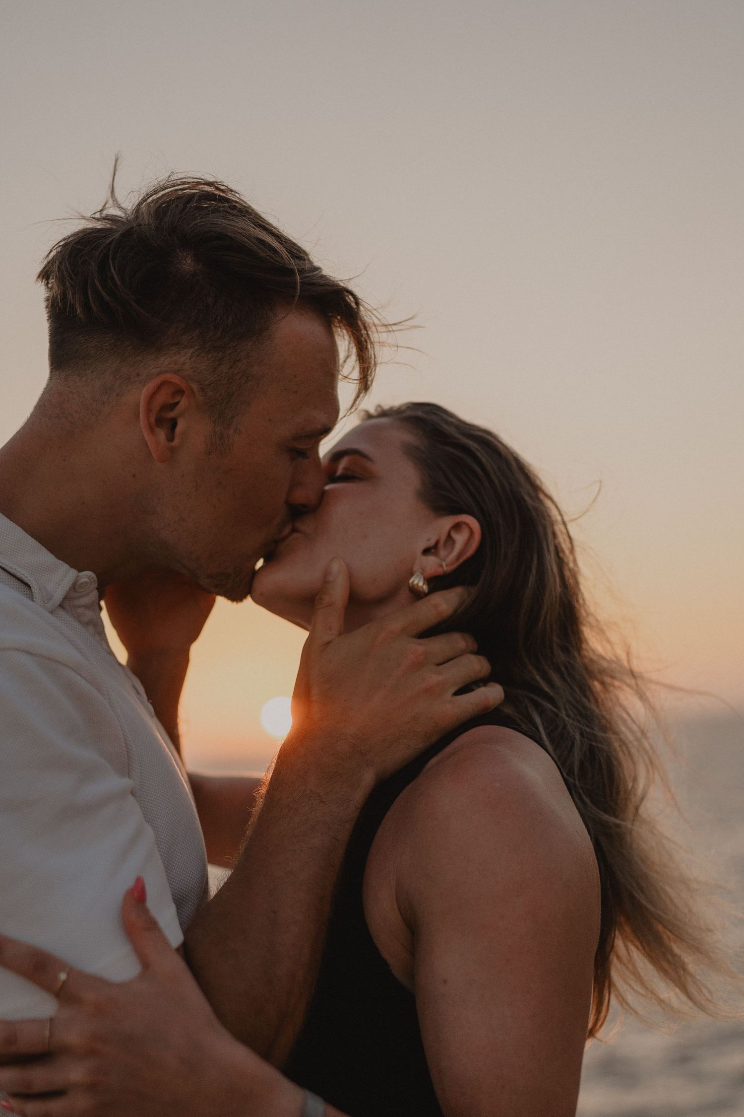 A couple shares a romantic kiss at sunset by the ocean, with warm golden light glowing behind them.