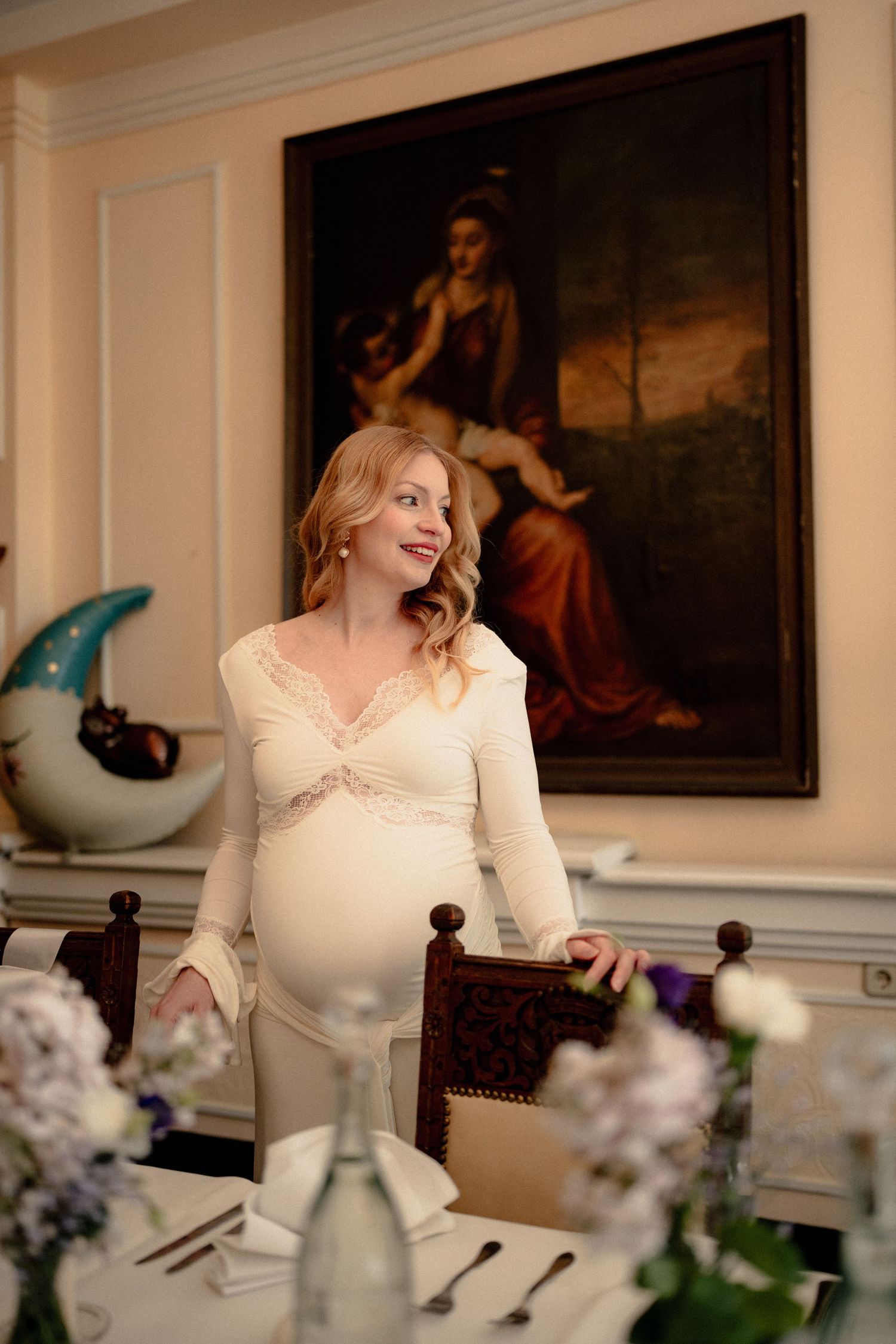 Smiling pregnant woman in cream lace dress standing by elegant dining table with classical painting on wall behind her.
