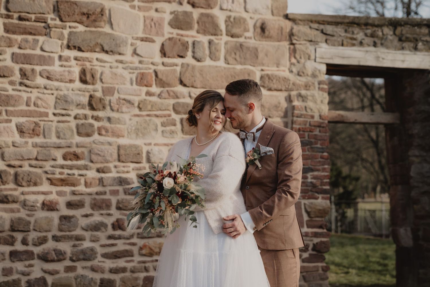 A couple shares an intimate moment against a rustic stone wall backdrop during their wedding celebration.