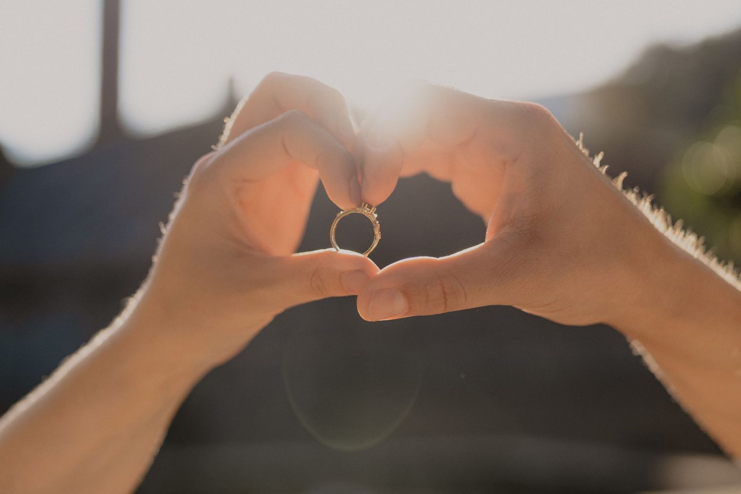 Hands forming a heart shape against sunlight with an engagement ring visible in silhouette.