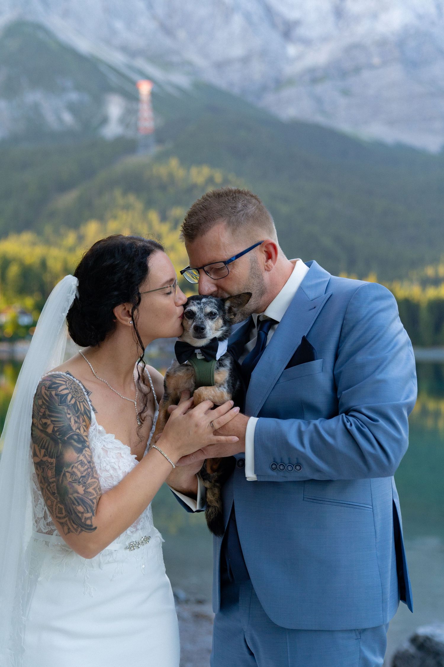 A couple kisses while holding their small dog during their wedding ceremony by a scenic mountain lake.