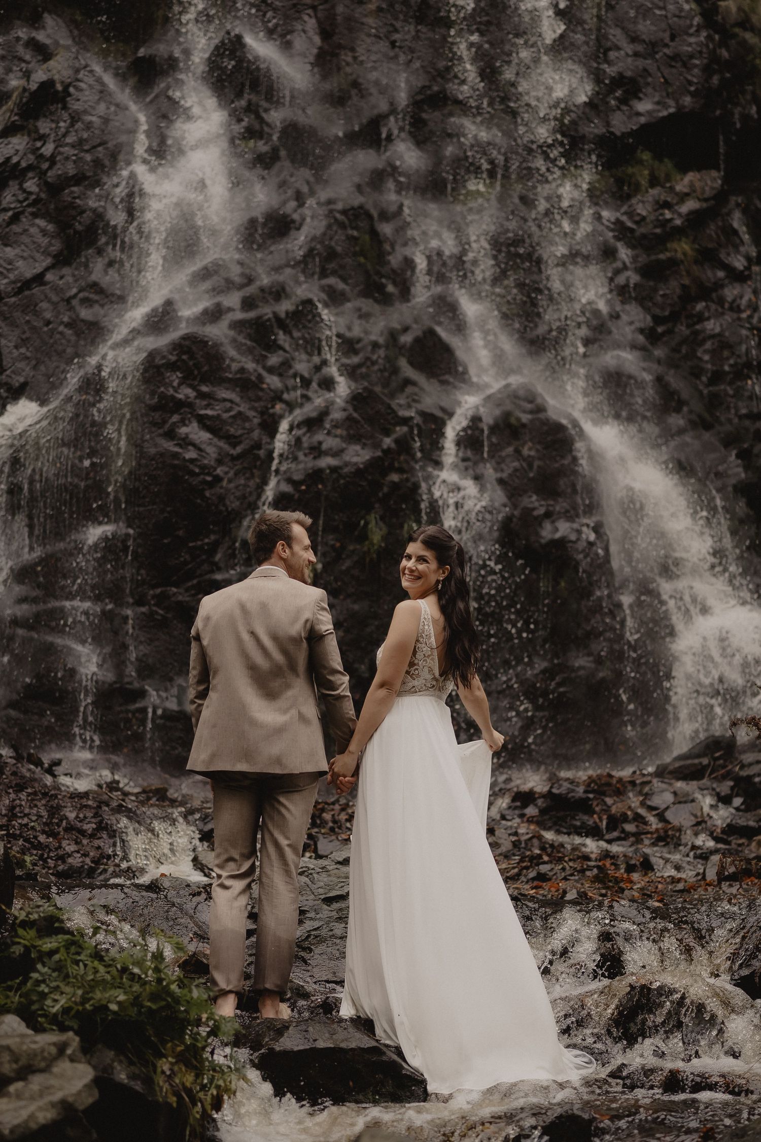 Newlyweds stand together against a misty waterfall in an intimate moment.