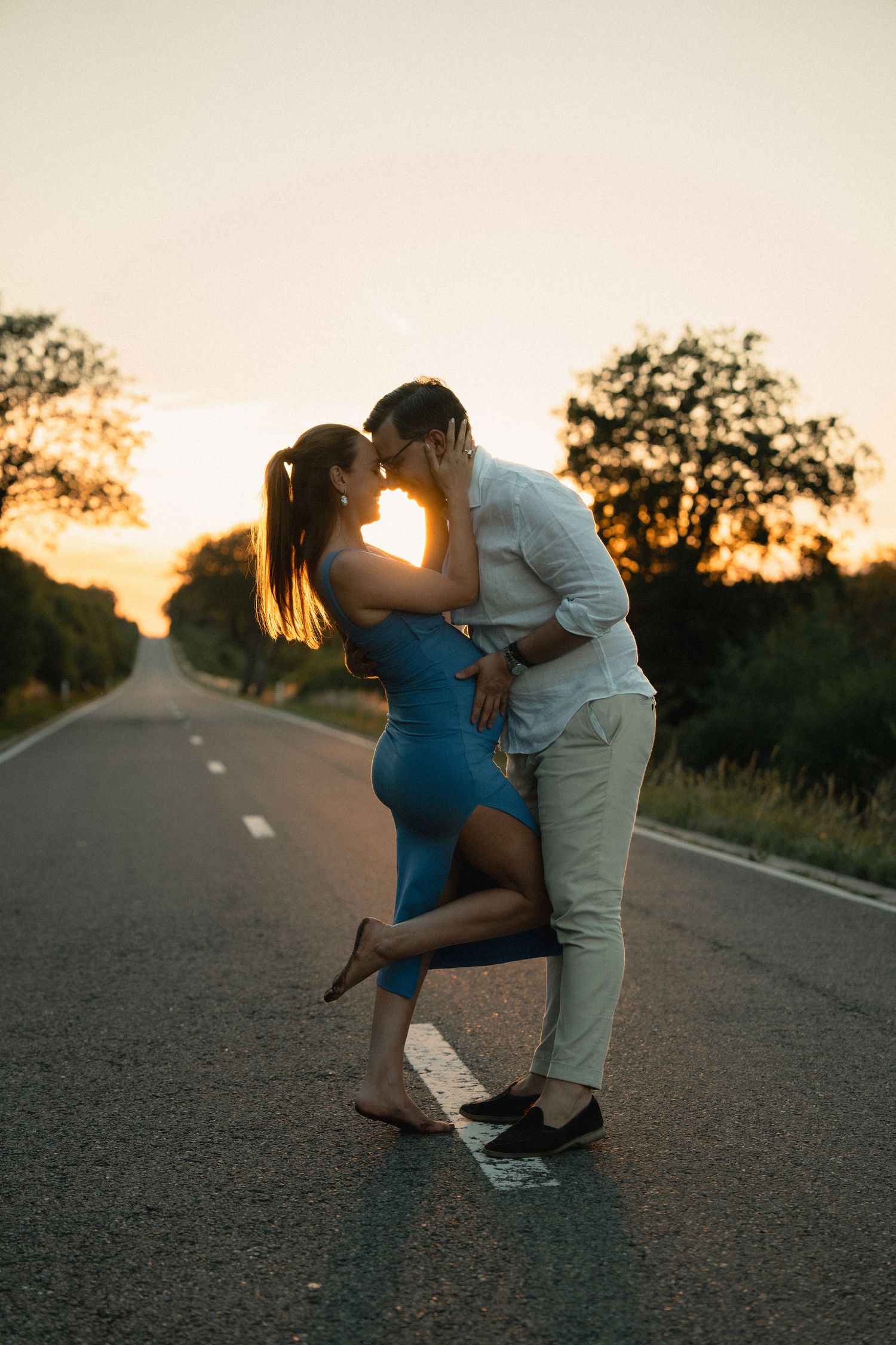Couple sharing a romantic kiss on an empty road at golden sunset, woman in blue dress lifted by man in white shirt.
