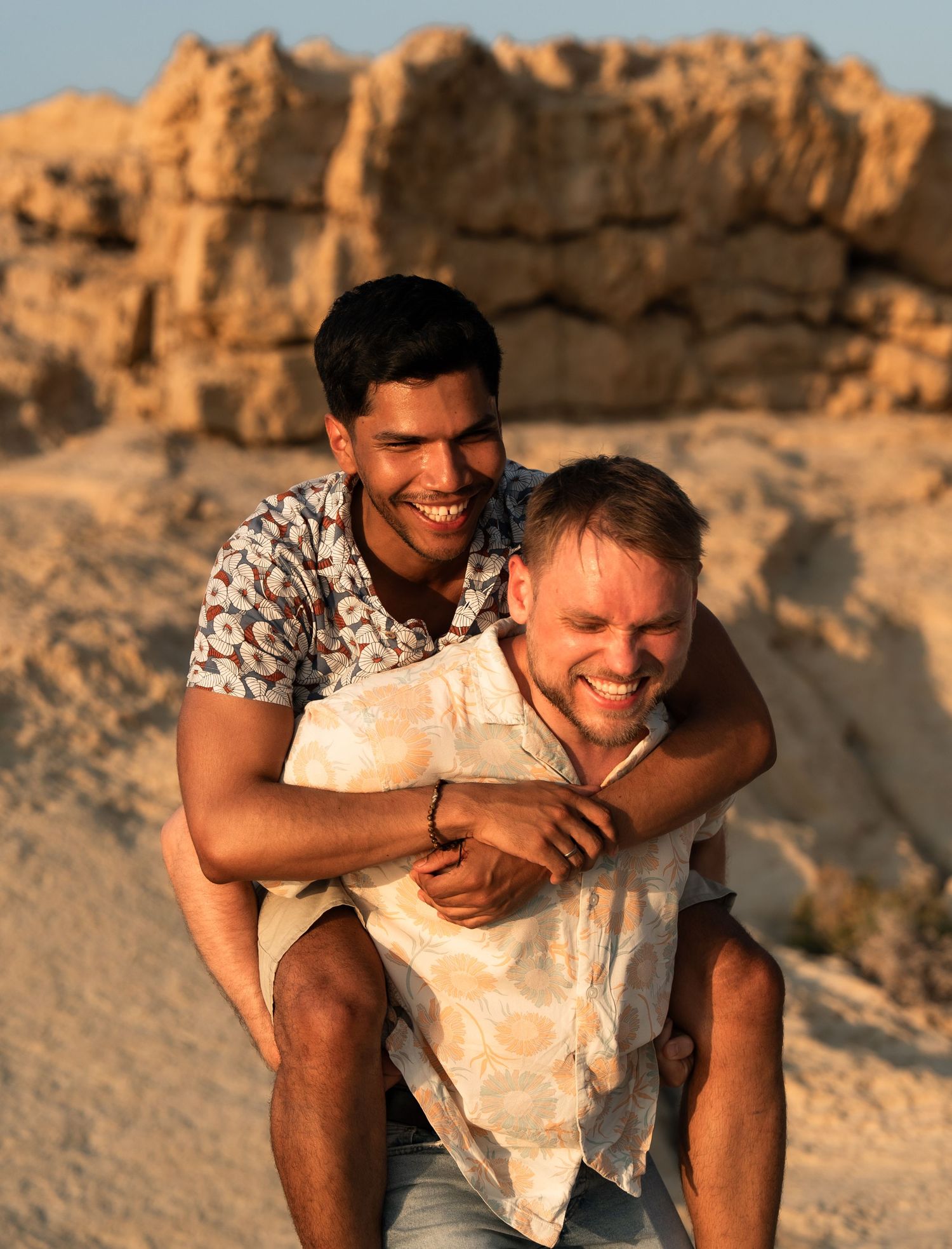 Two smiling men enjoy a piggyback ride on sandy desert dunes at golden hour.