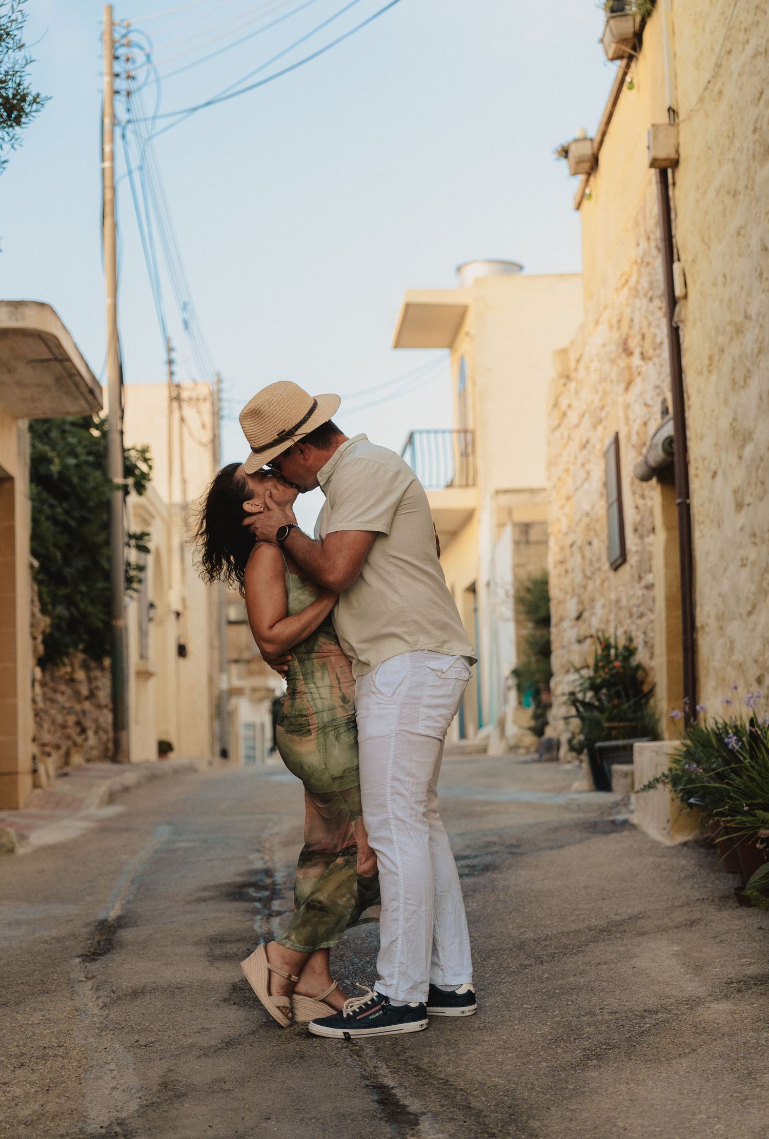 Couple sharing a romantic kiss on a charming Mediterranean village street, man in white pants and hat, woman in green outfit.