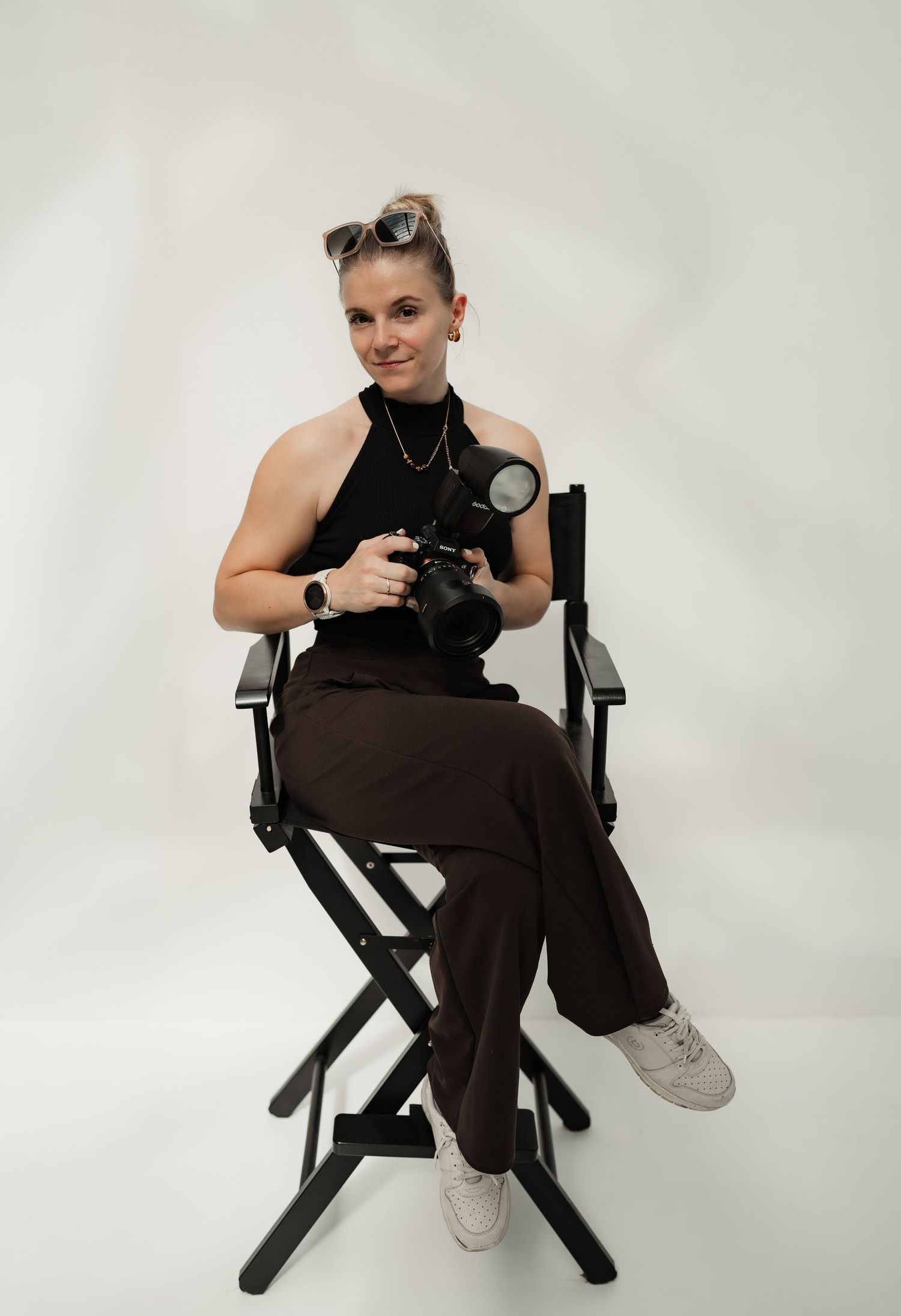 Female photographer sitting in director's chair holding camera with flash, wearing black top and brown pants.