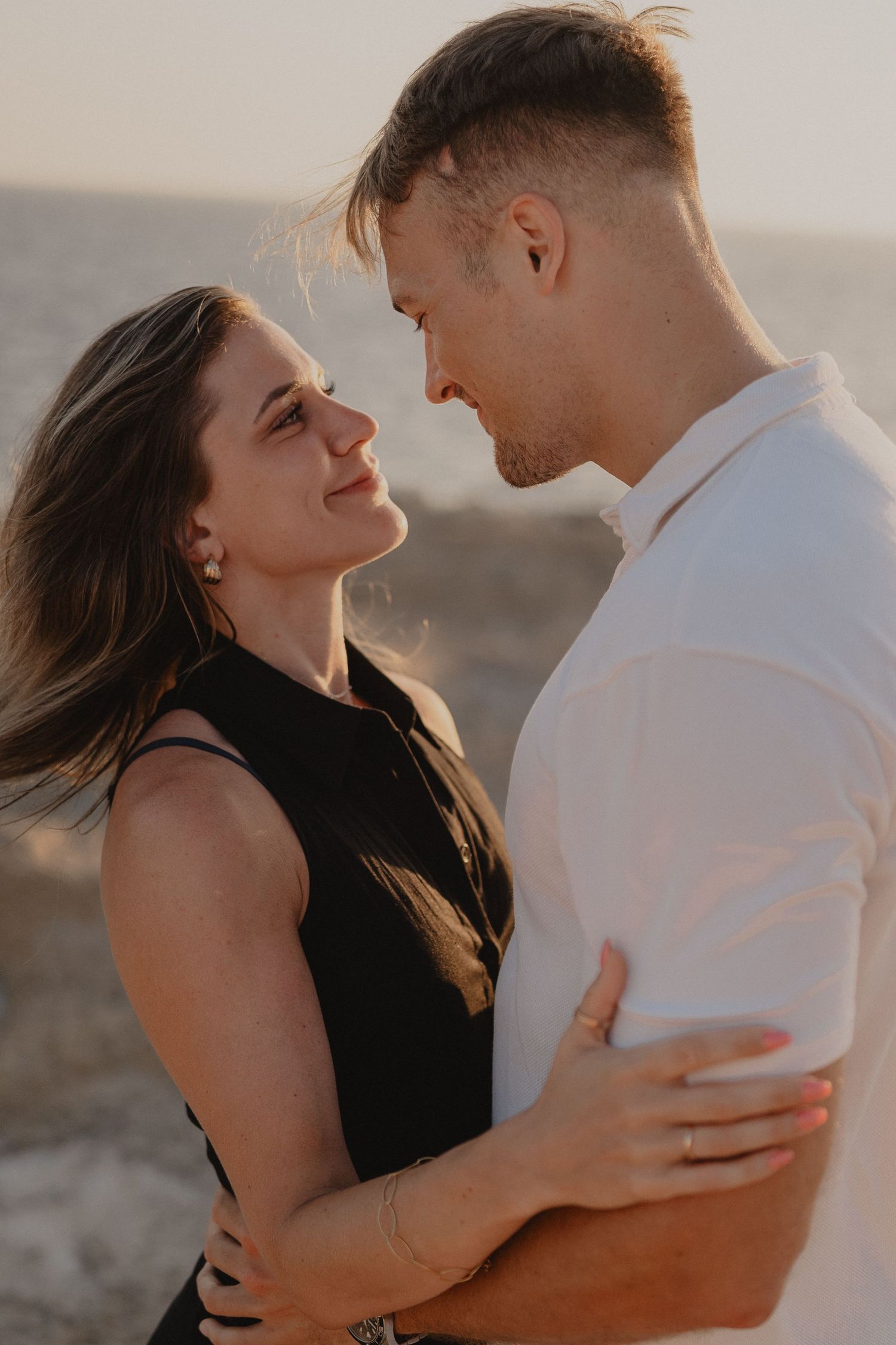 A couple embraces romantically at sunset, the woman in black gazing up at the man in white against a scenic backdrop.