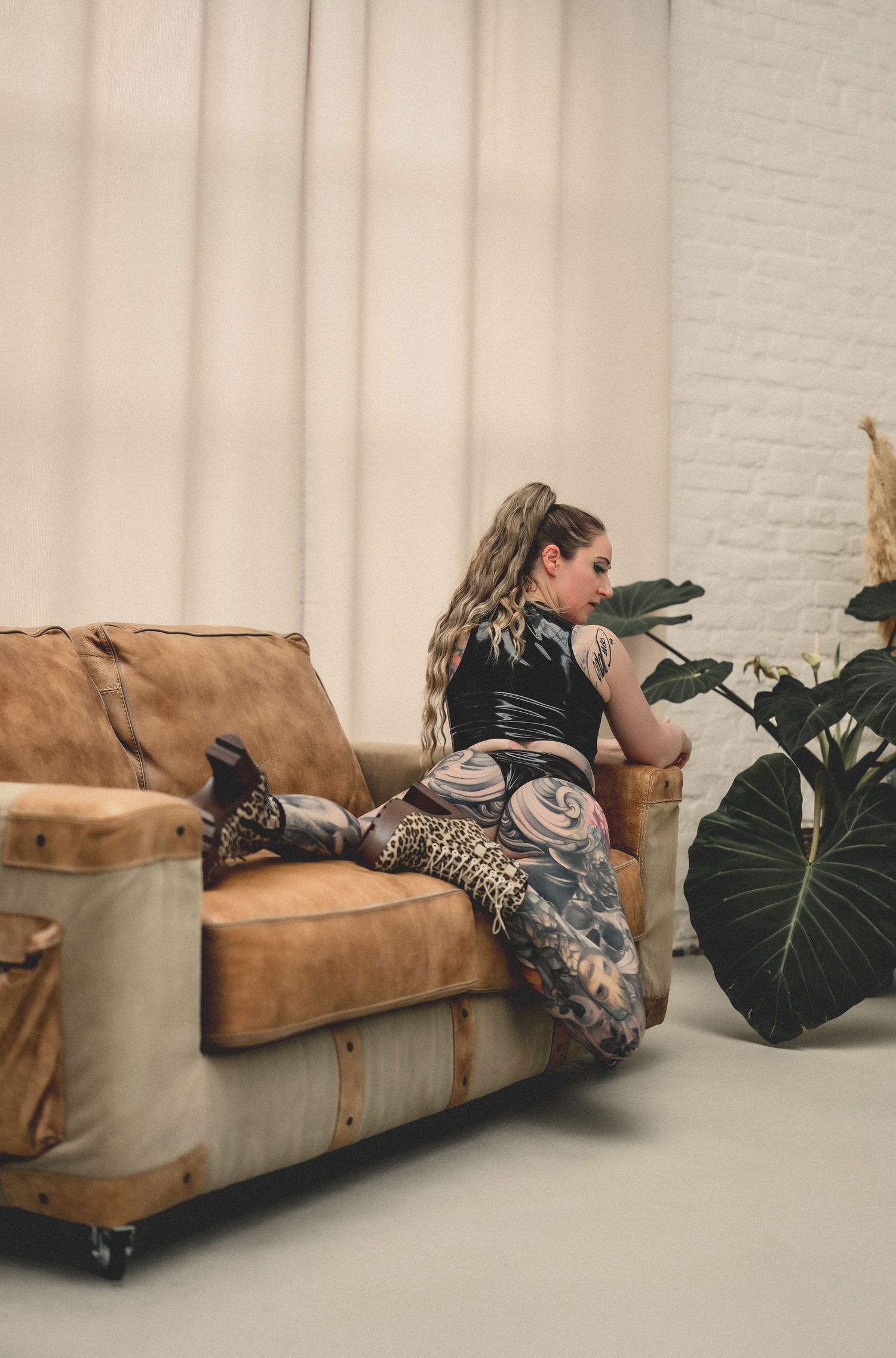 Tattooed woman in patterned pants sitting on vintage leather sofa near large plant and white curtains.