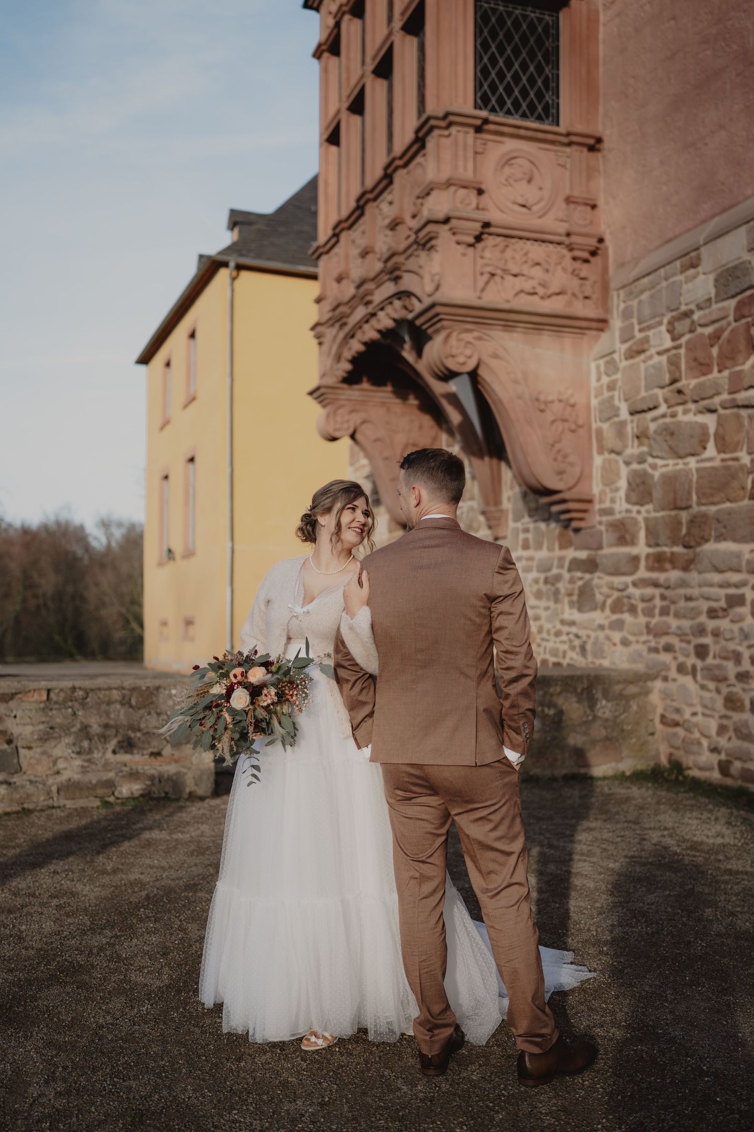 A romantic wedding couple walking together outside a historic stone building in brown and white attire.