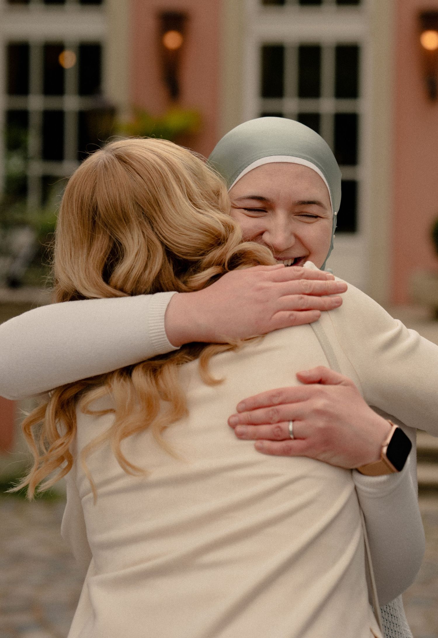 Two women share a warm embrace at a baby shower, one wearing a sage green hijab and a smartwatch.