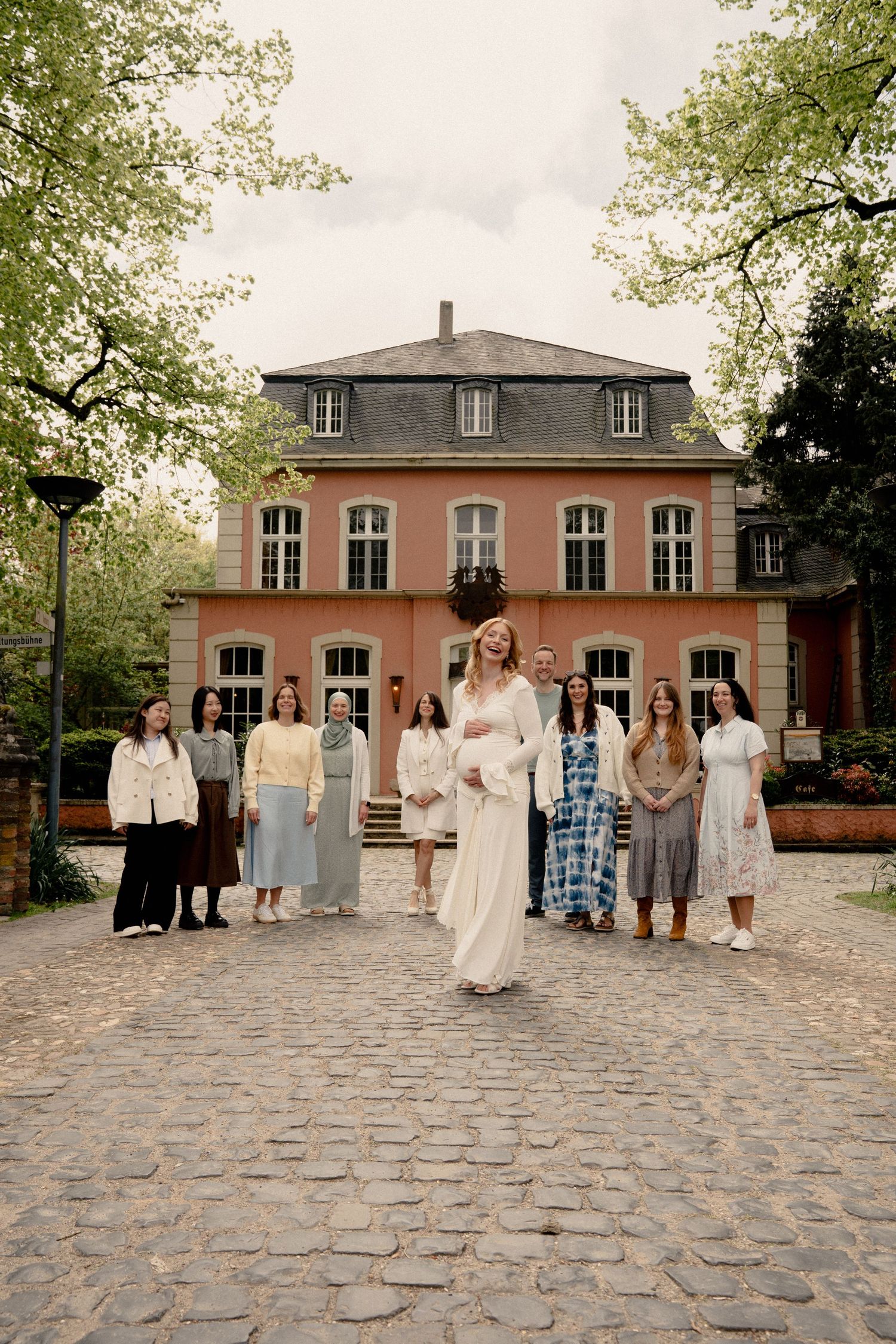 Group of women in elegant dresses posing in front of a pink historic building on a cobblestone courtyard.