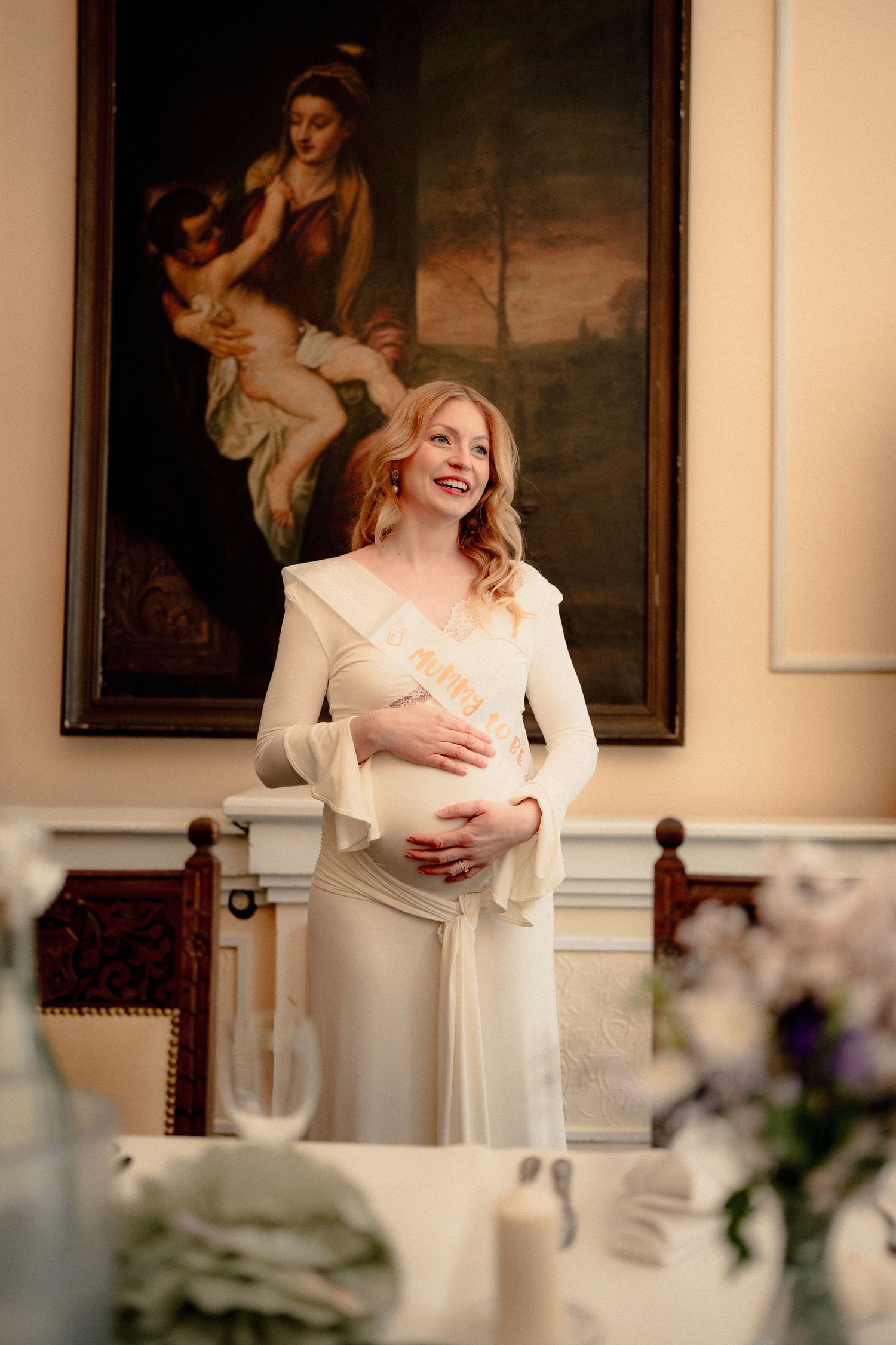Elegant pregnant woman wearing mama sash standing in ornate room with classical painting.