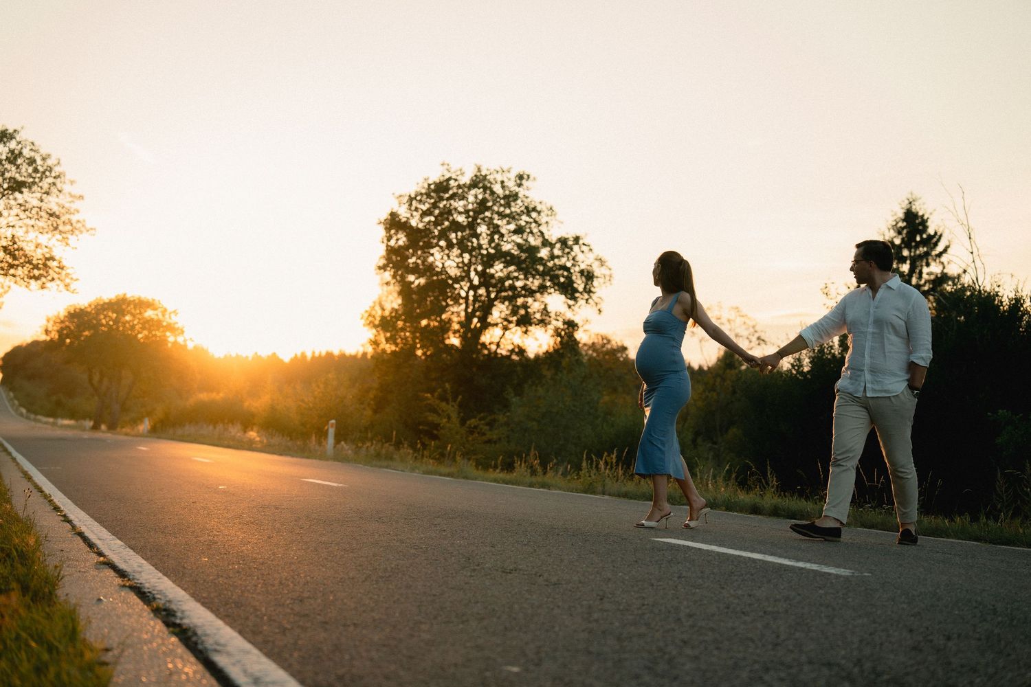 Couple holding hands walking on a road at sunset with golden light and trees in the background.