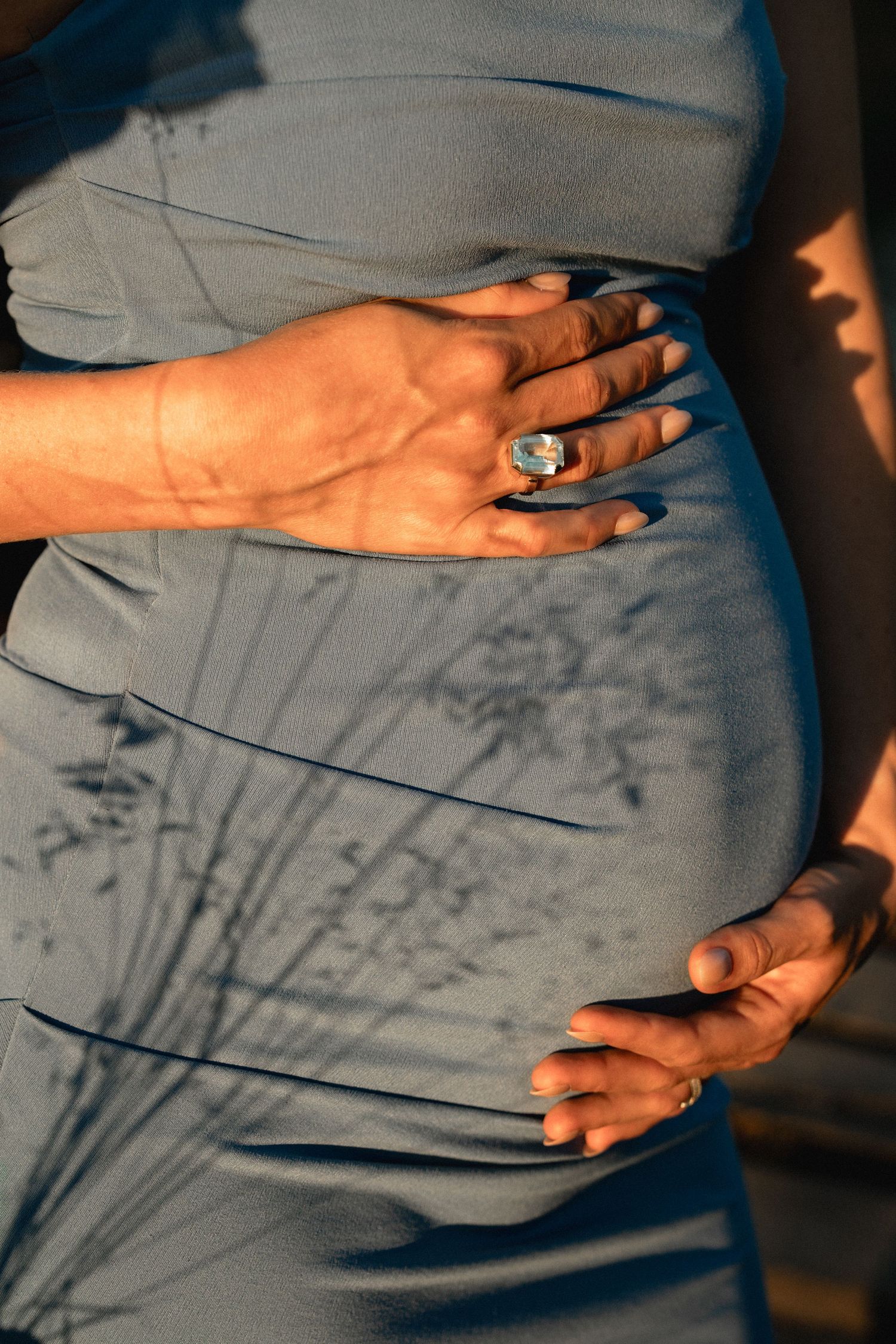 Close-up of pregnant woman in blue dress cradling baby bump, wearing a large gemstone ring in warm sunlight.