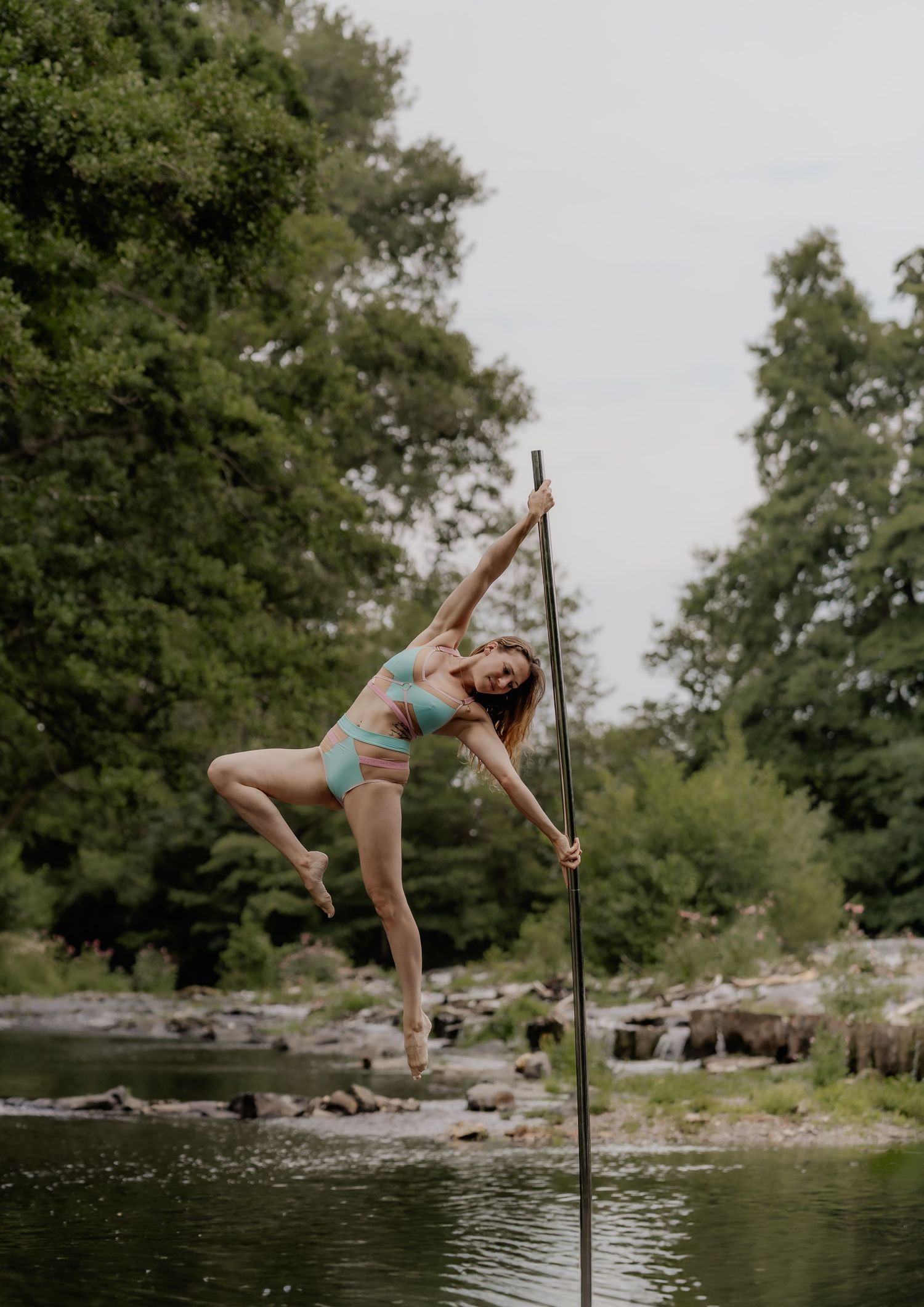 A woman in a bikini performs an aerial pole dance move outdoors near a river surrounded by lush green trees.