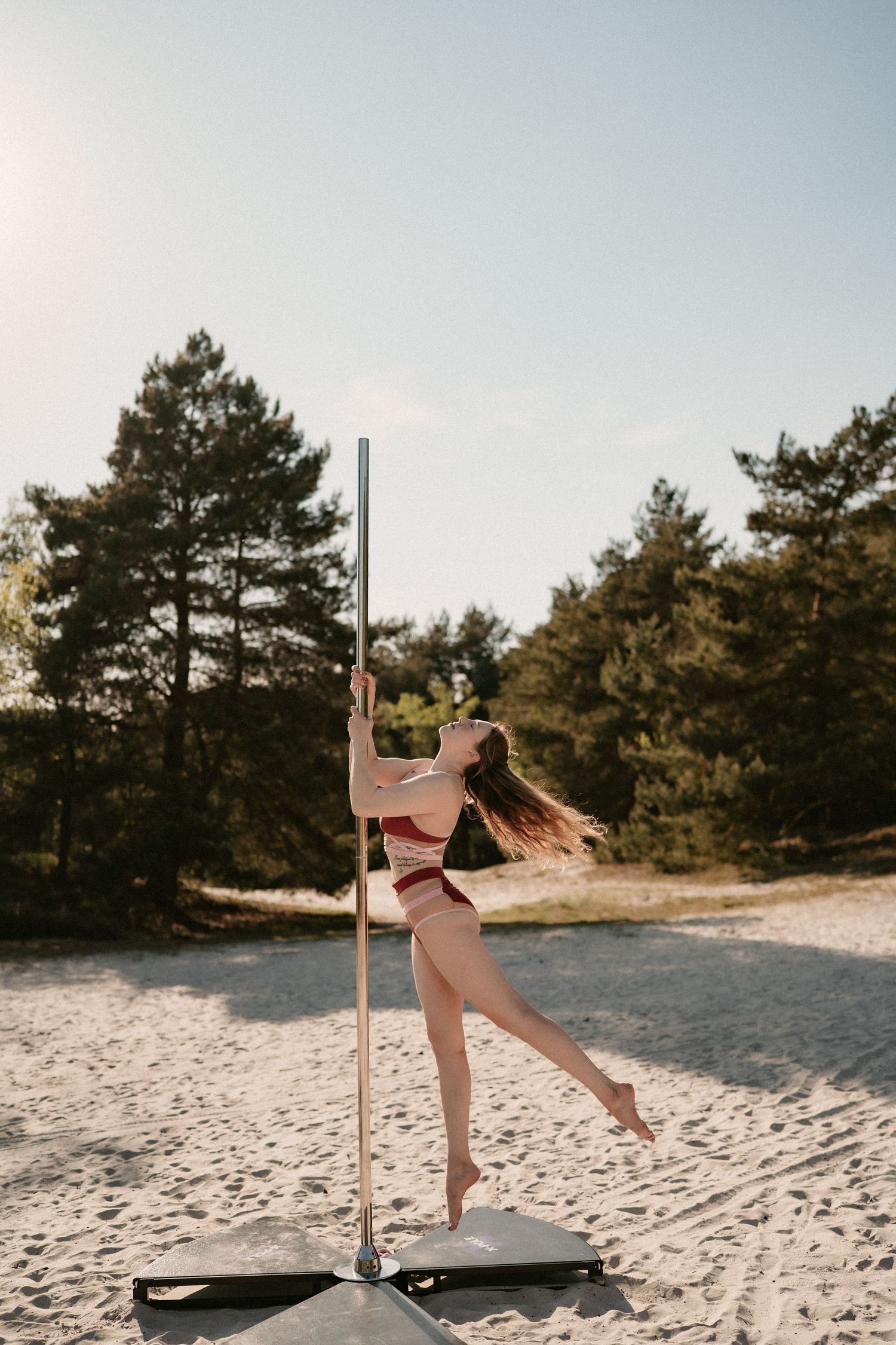 A woman in a red bikini performs pole dancing on a sandy beach surrounded by pine trees on a sunny day.