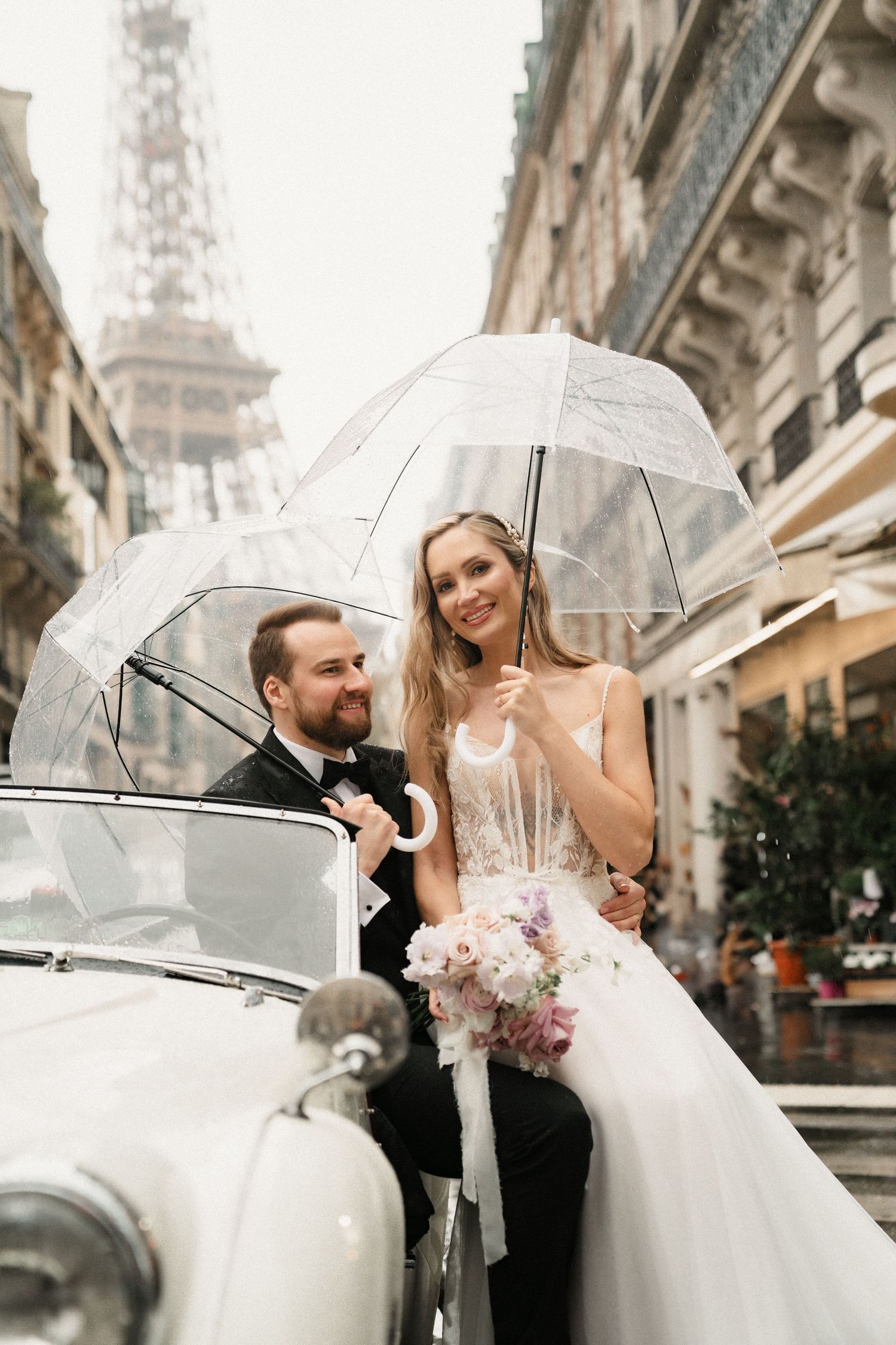 Romantic wedding couple shares clear umbrella in front of Eiffel Tower on rainy Paris day with vintage car.