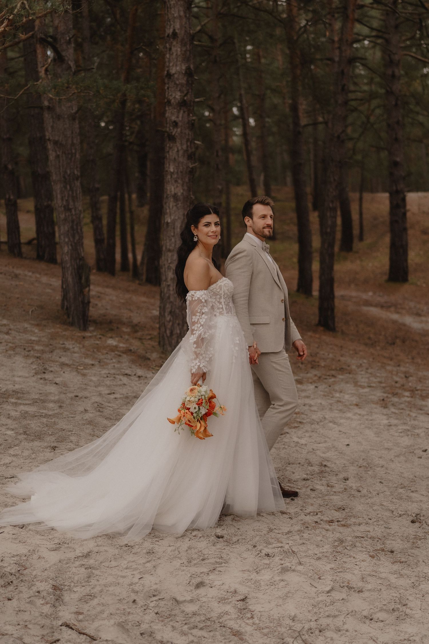 Wedding couple poses in a pine forest with sandy ground and autumn-colored bouquet.