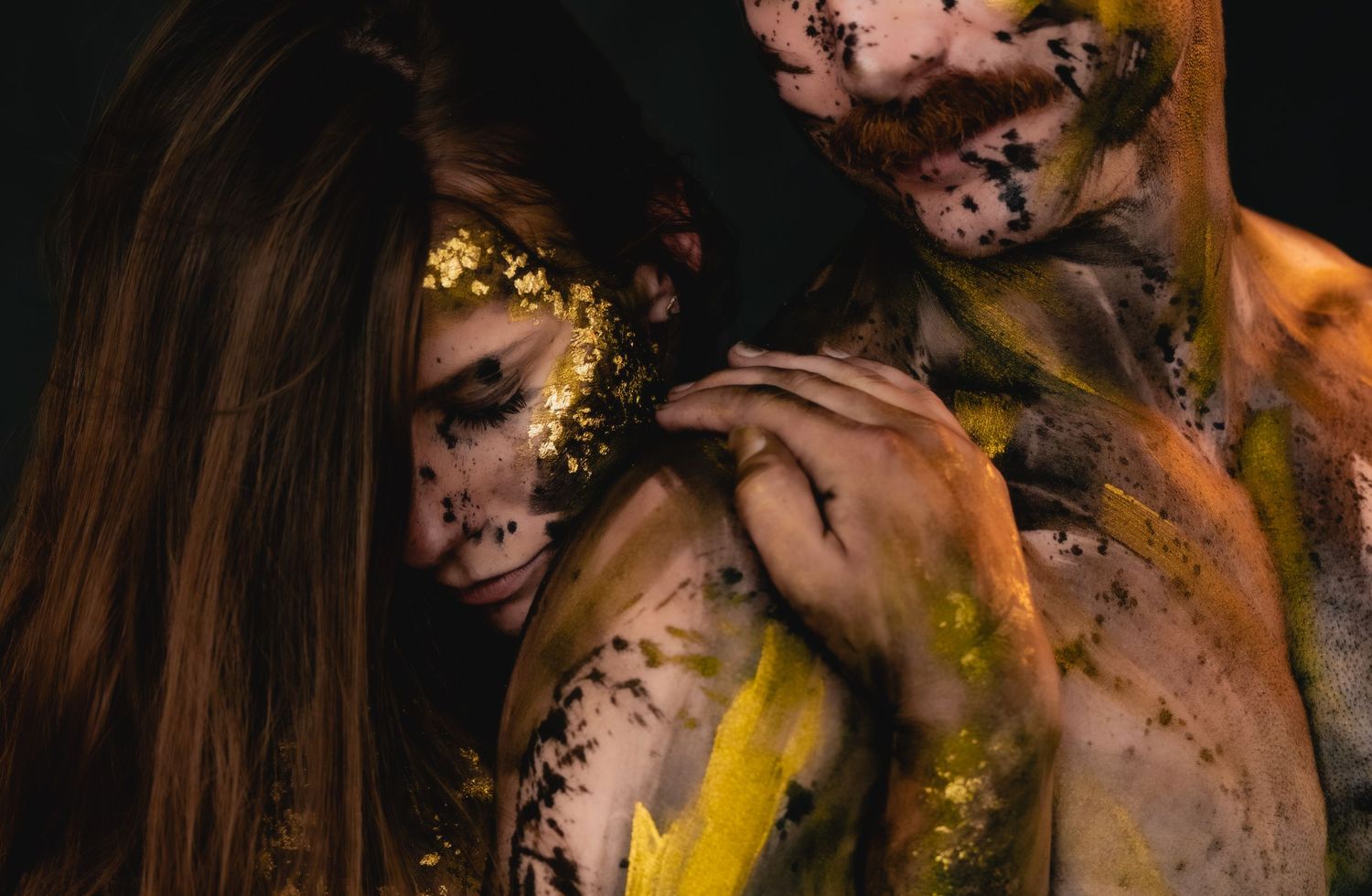Two people with artistic body paint featuring gold glitter and yellow pigments pose dramatically against a dark background.
