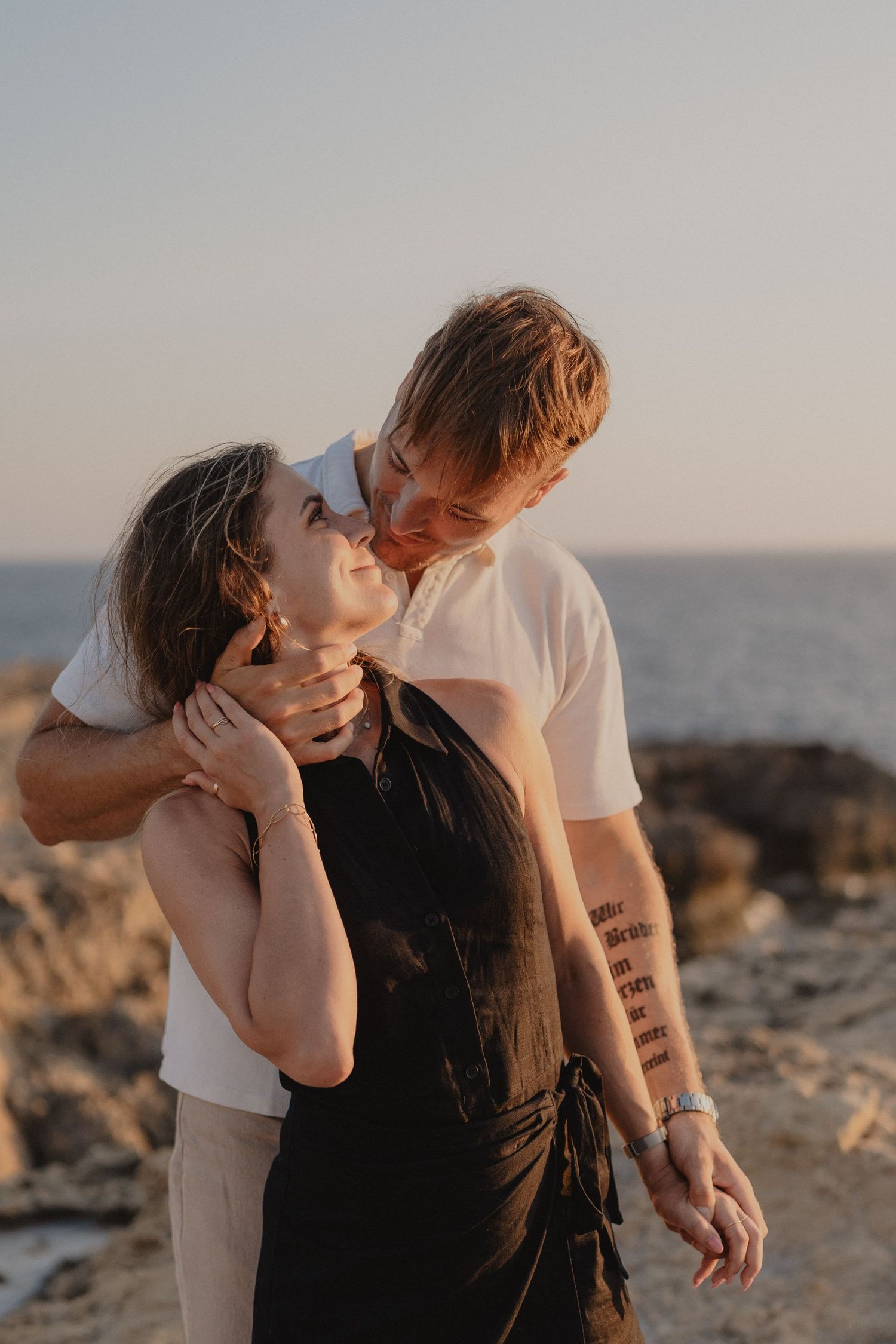 Couple embracing at sunset on rocky coast, man kissing woman's cheek, ocean backdrop.