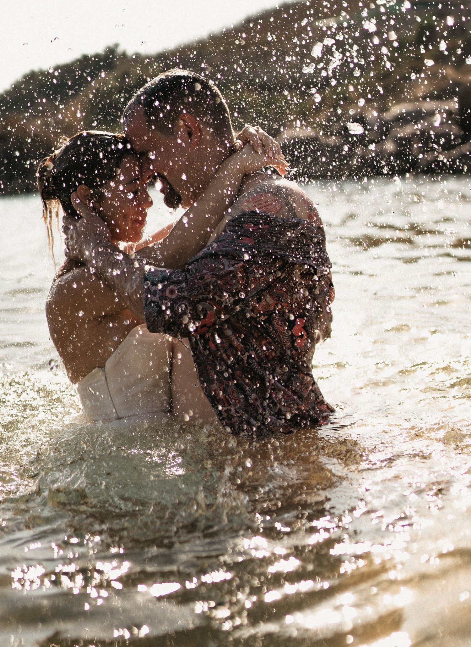Water splashes around a couple during a playful moment in the ocean at sunset.