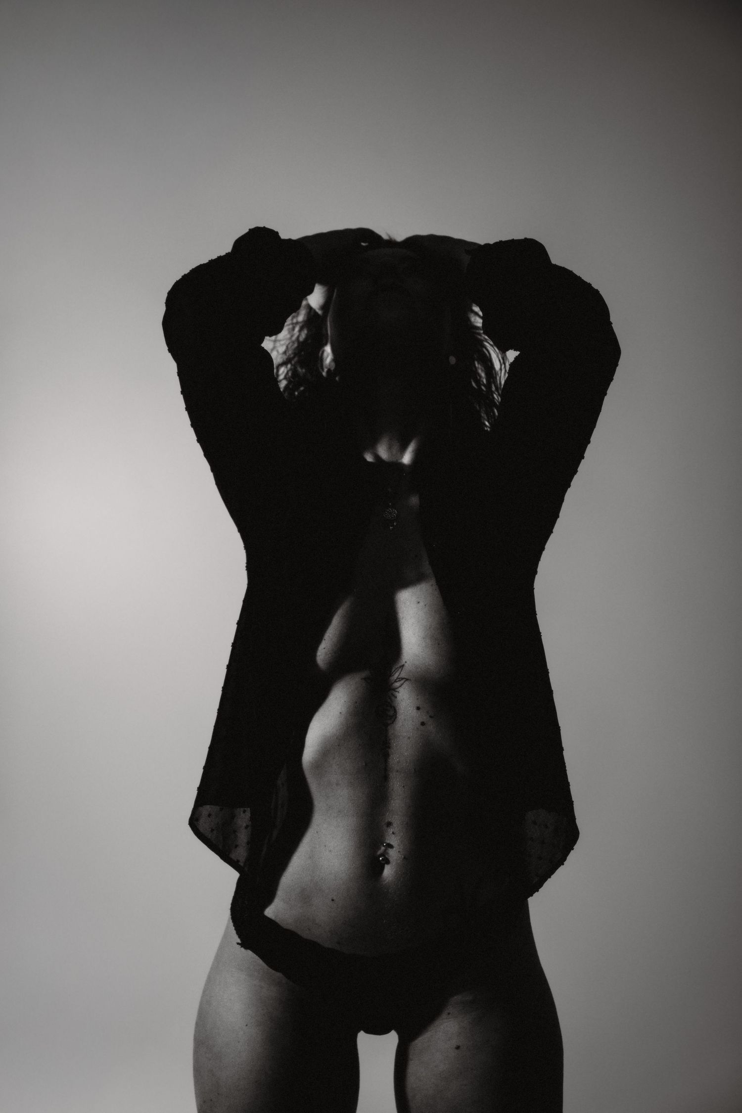 Black and white artistic photo of a woman removing a dark top, showing torso in dim studio lighting.