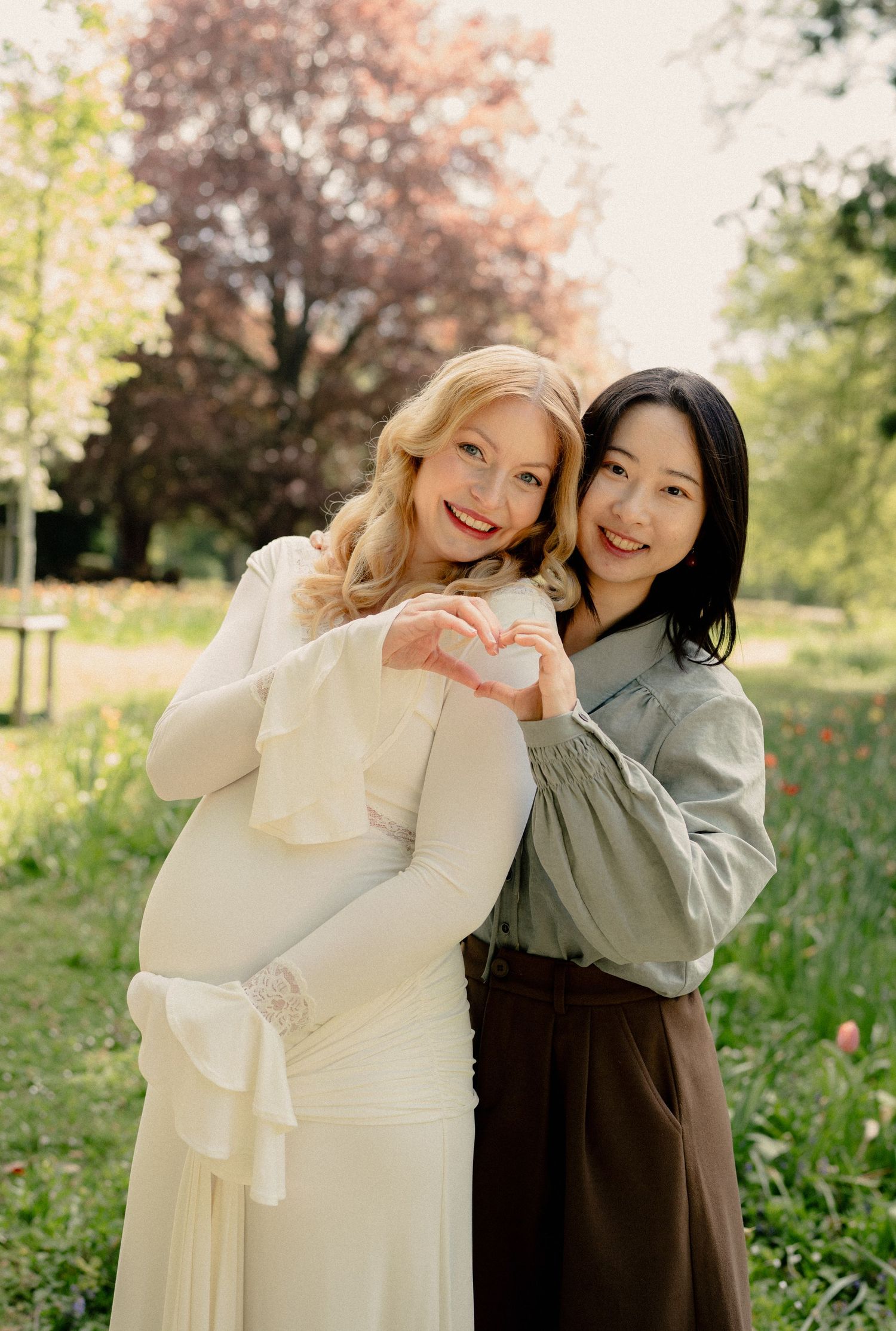 Two women making heart shape with hands in sunny garden, one visibly pregnant in white dress.