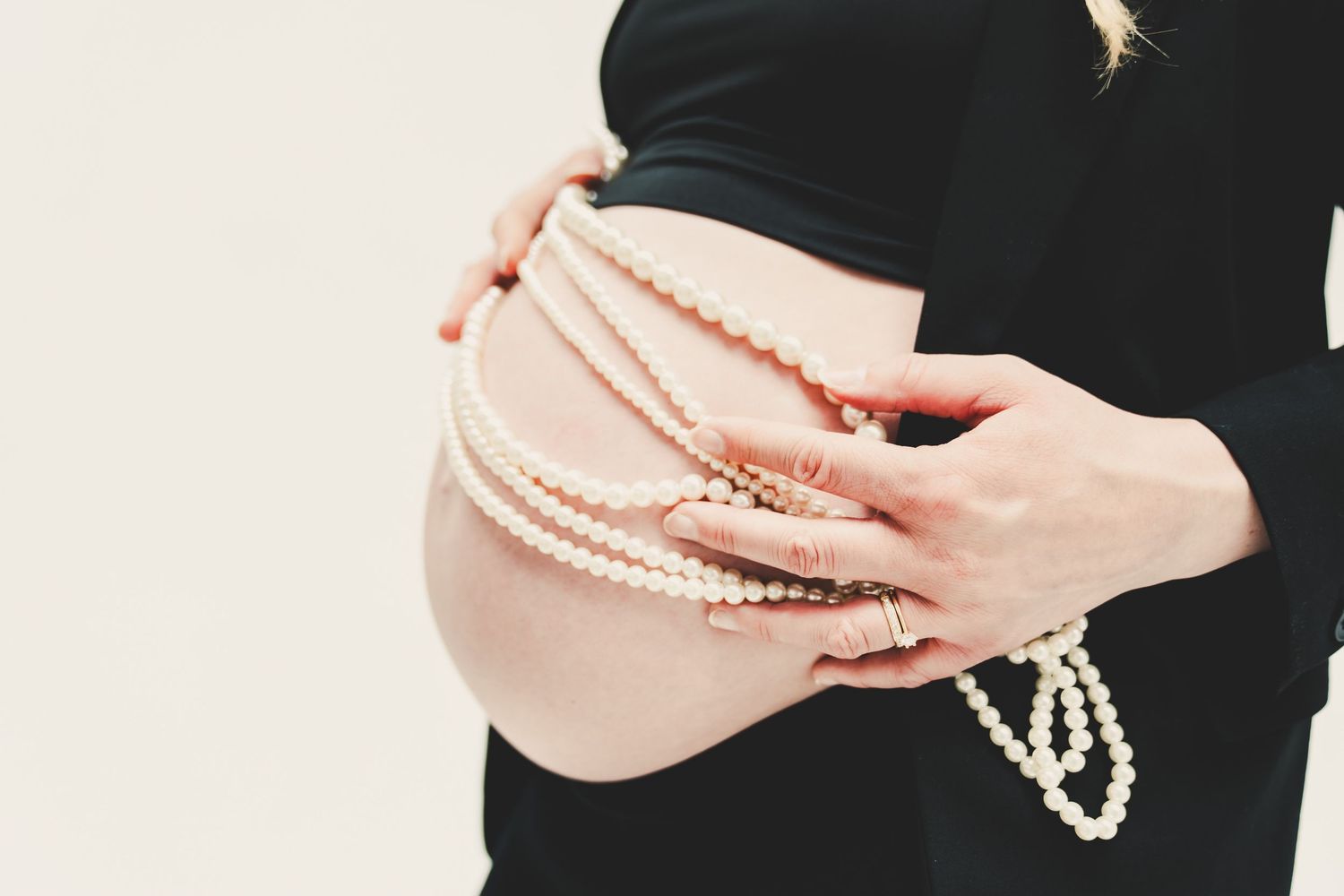 Pregnant woman in black dress cradling bare baby bump with hands adorned in pearl necklaces and gold rings.