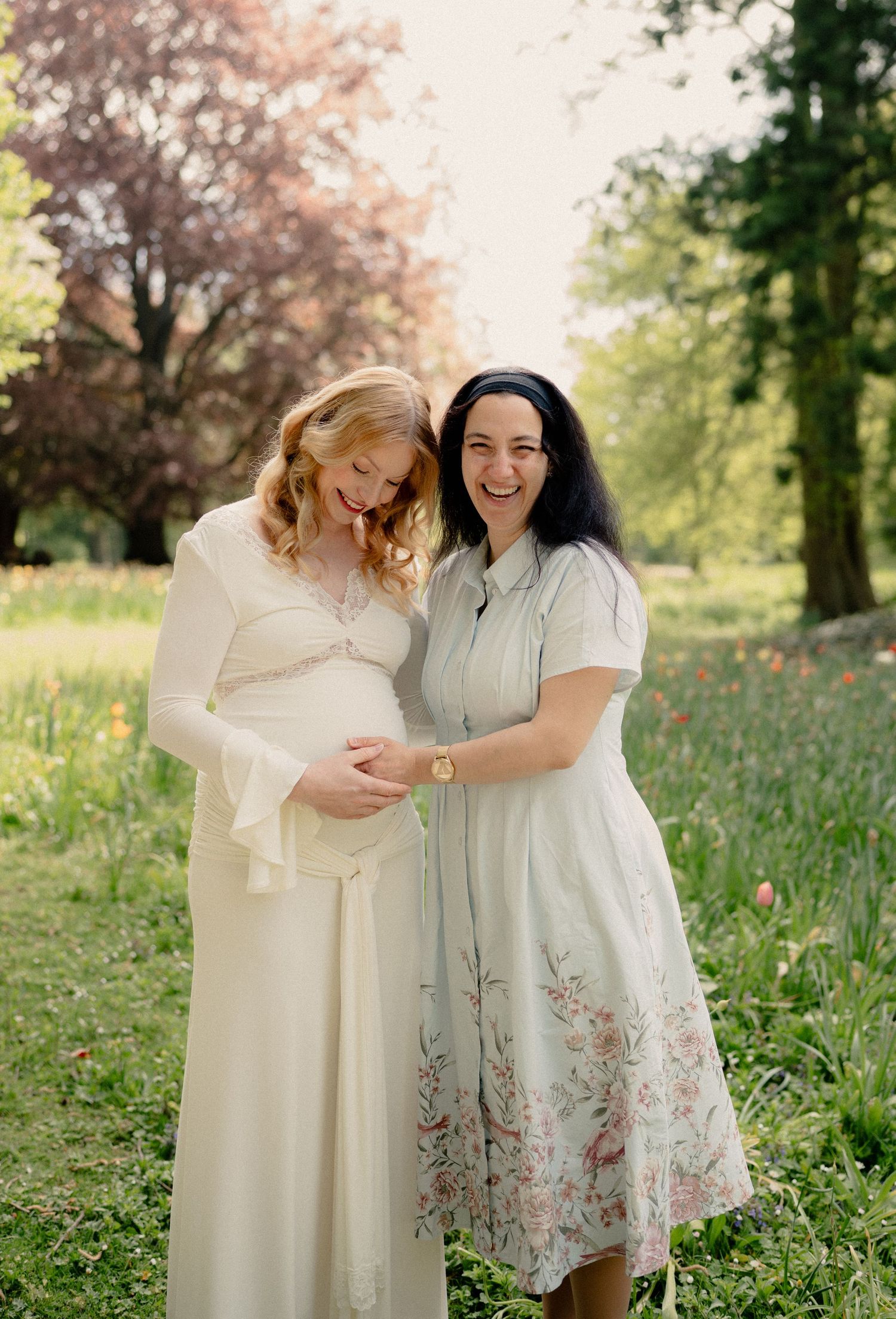 Laughing pregnant woman in cream gown poses with dark-haired friend in floral dress in a blooming spring garden.
