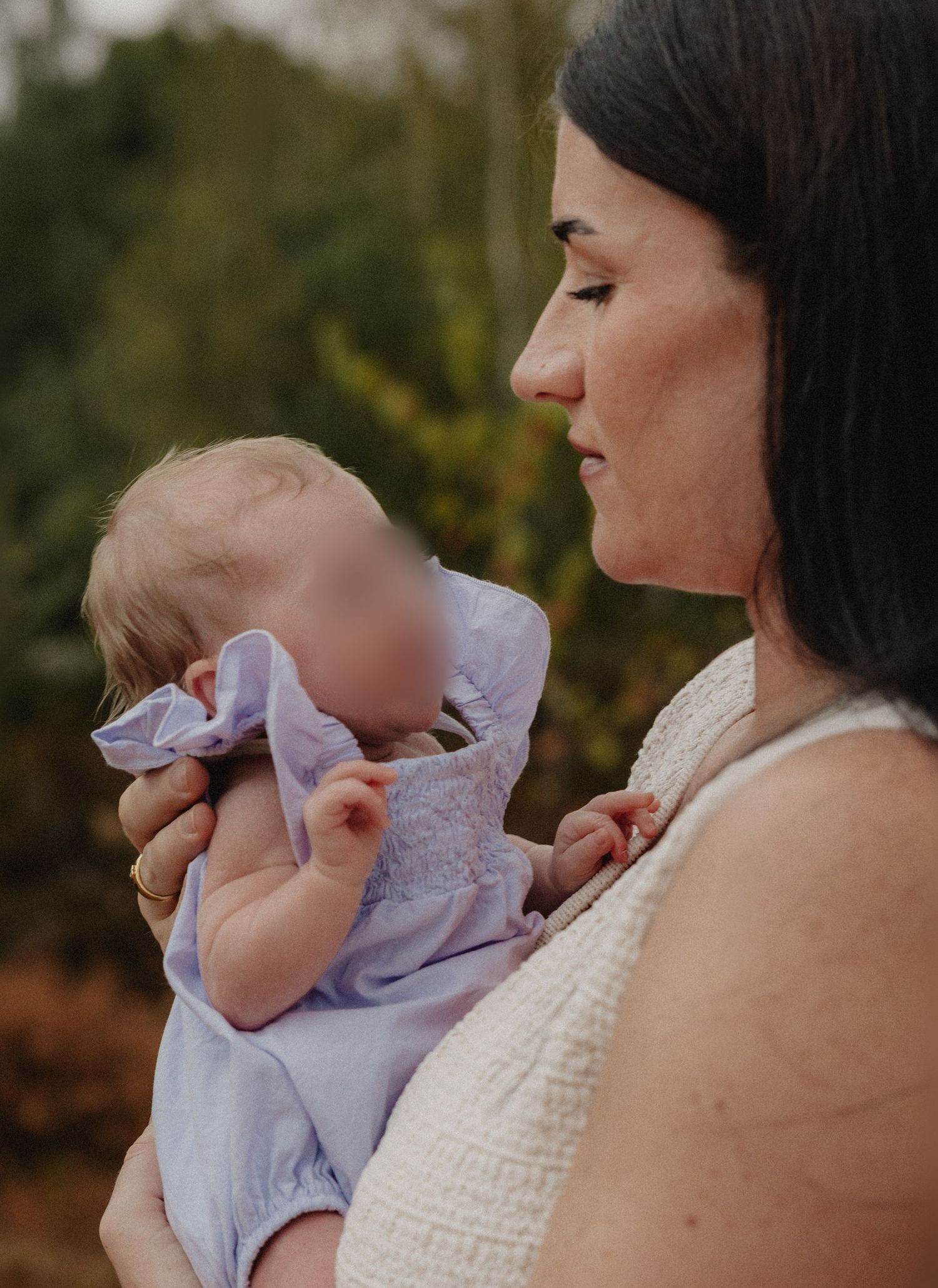 A dark-haired woman tenderly holds a baby dressed in lavender outdoors with green trees in the background.