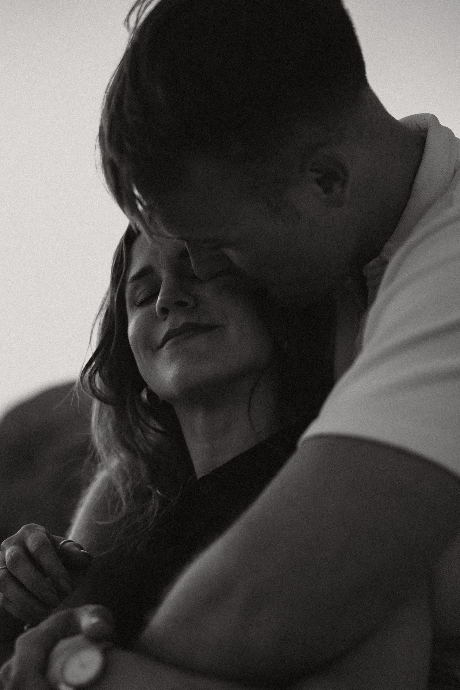 Black and white photo of a couple embracing tenderly, man resting his head on womans head while holding hands.