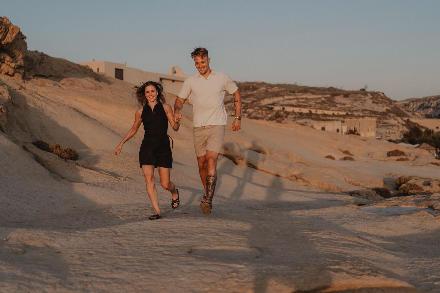 Couple holding hands walking on sandy rocky terrain at golden hour sunset in a desert landscape.