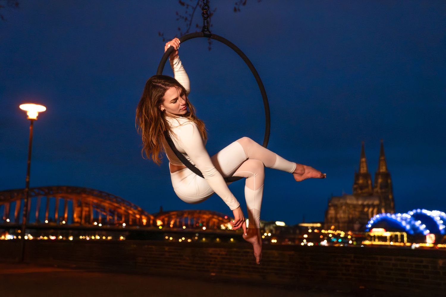 Aerial acrobat performs on a hoop ring at night with Cologne Cathedral and bridge lights in background.